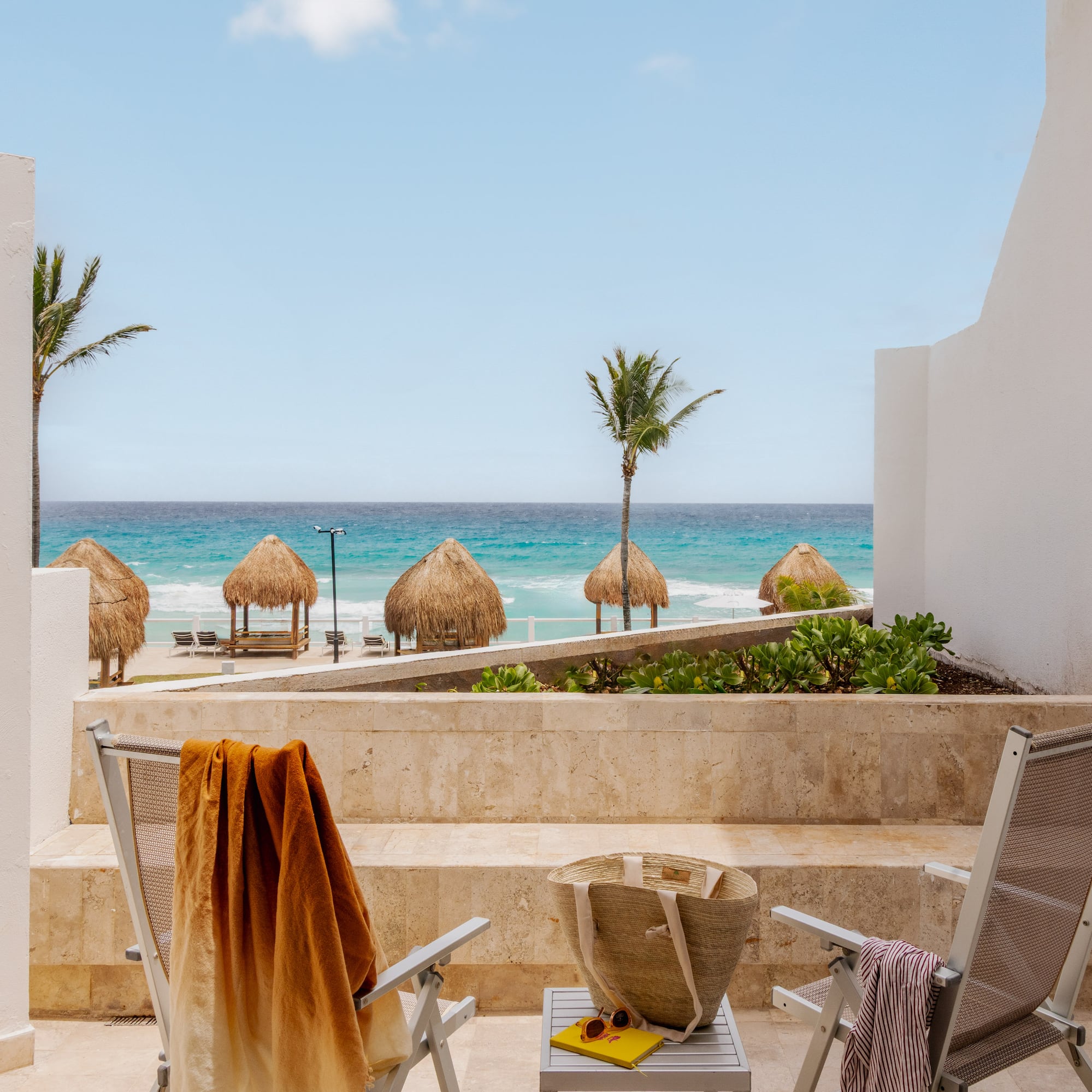 Oceanfront balcony view of tropical beach with palapas, palm trees, and chairs.