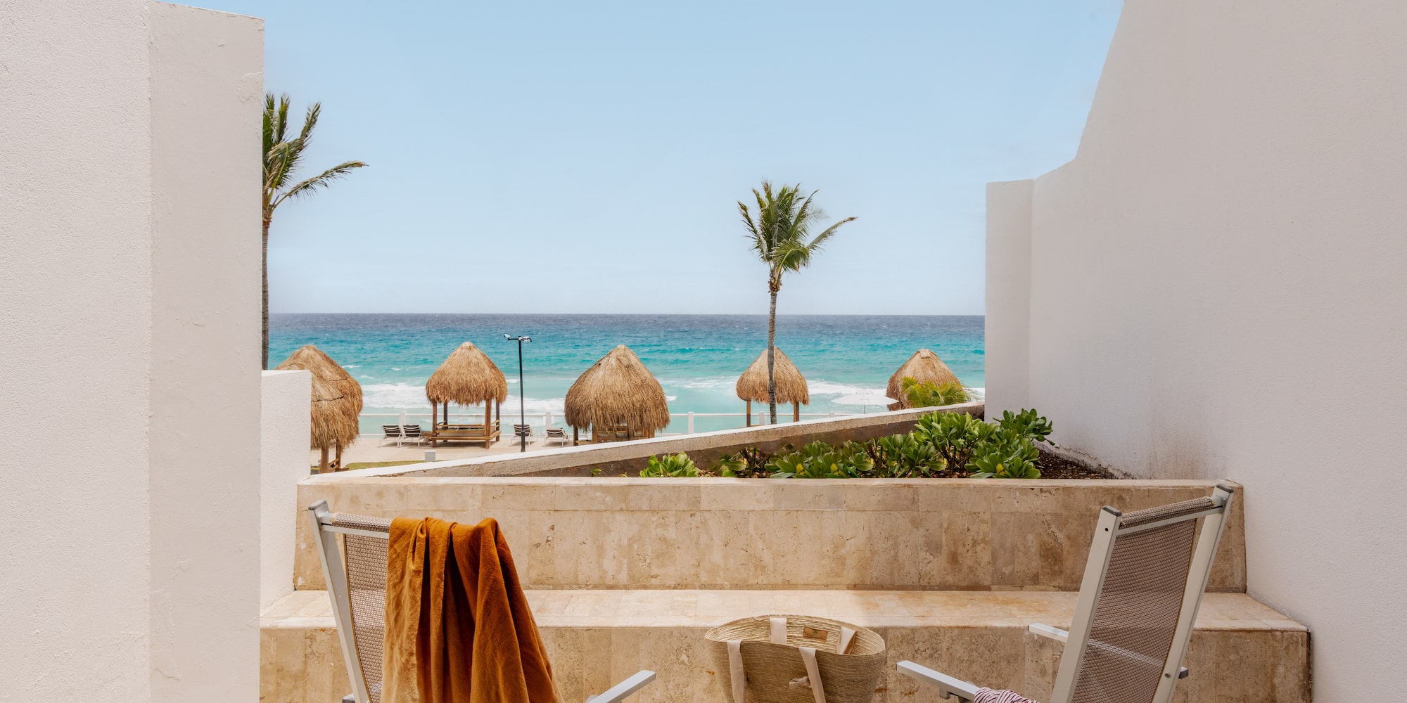 Oceanfront balcony view of tropical beach with palapas, palm trees, and chairs.