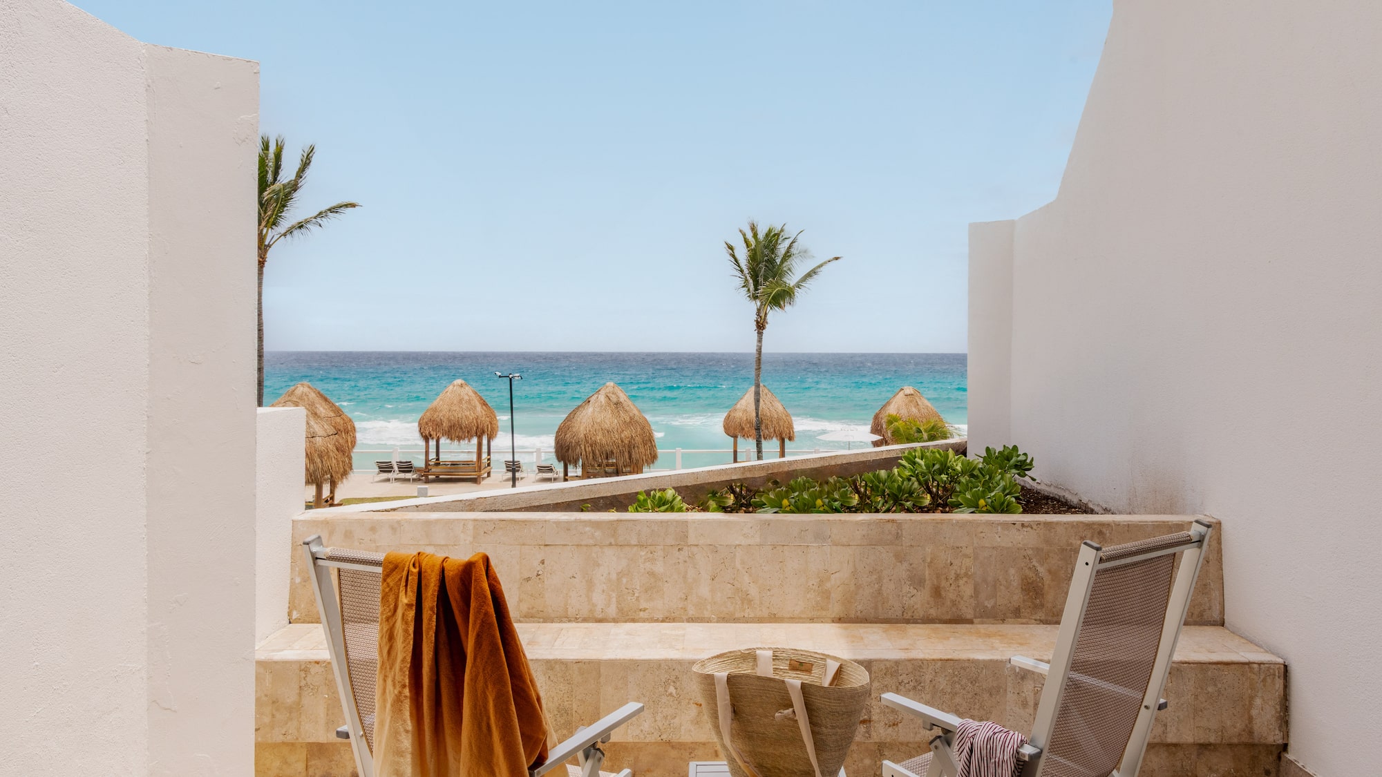 Oceanfront balcony view of tropical beach with palapas, palm trees, and chairs.