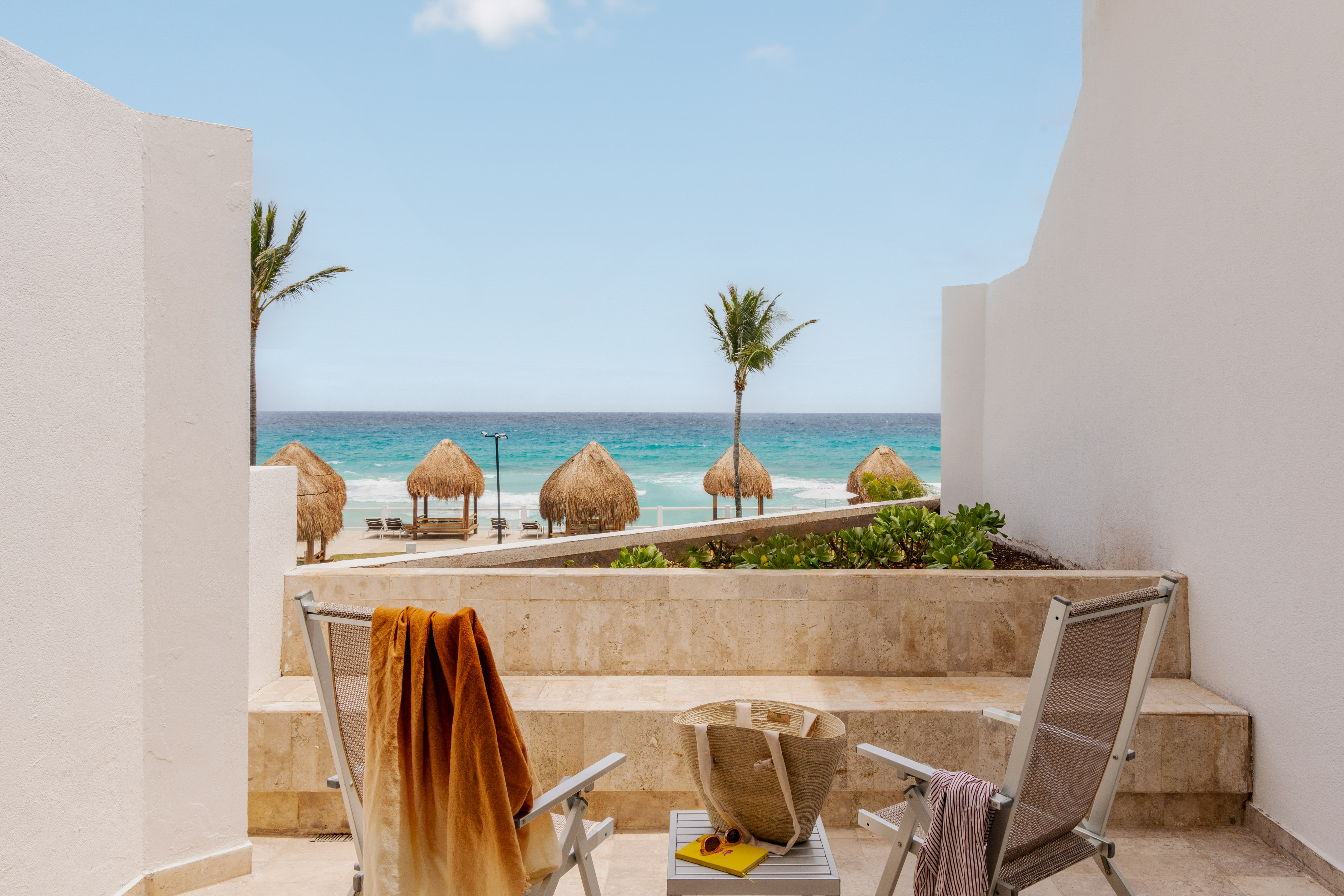 Oceanfront balcony view of tropical beach with palapas, palm trees, and chairs.