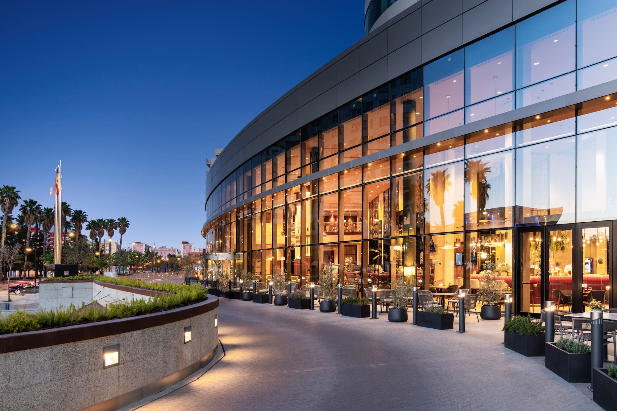 Modern curved glass hotel exterior at twilight with outdoor seating and palm trees.