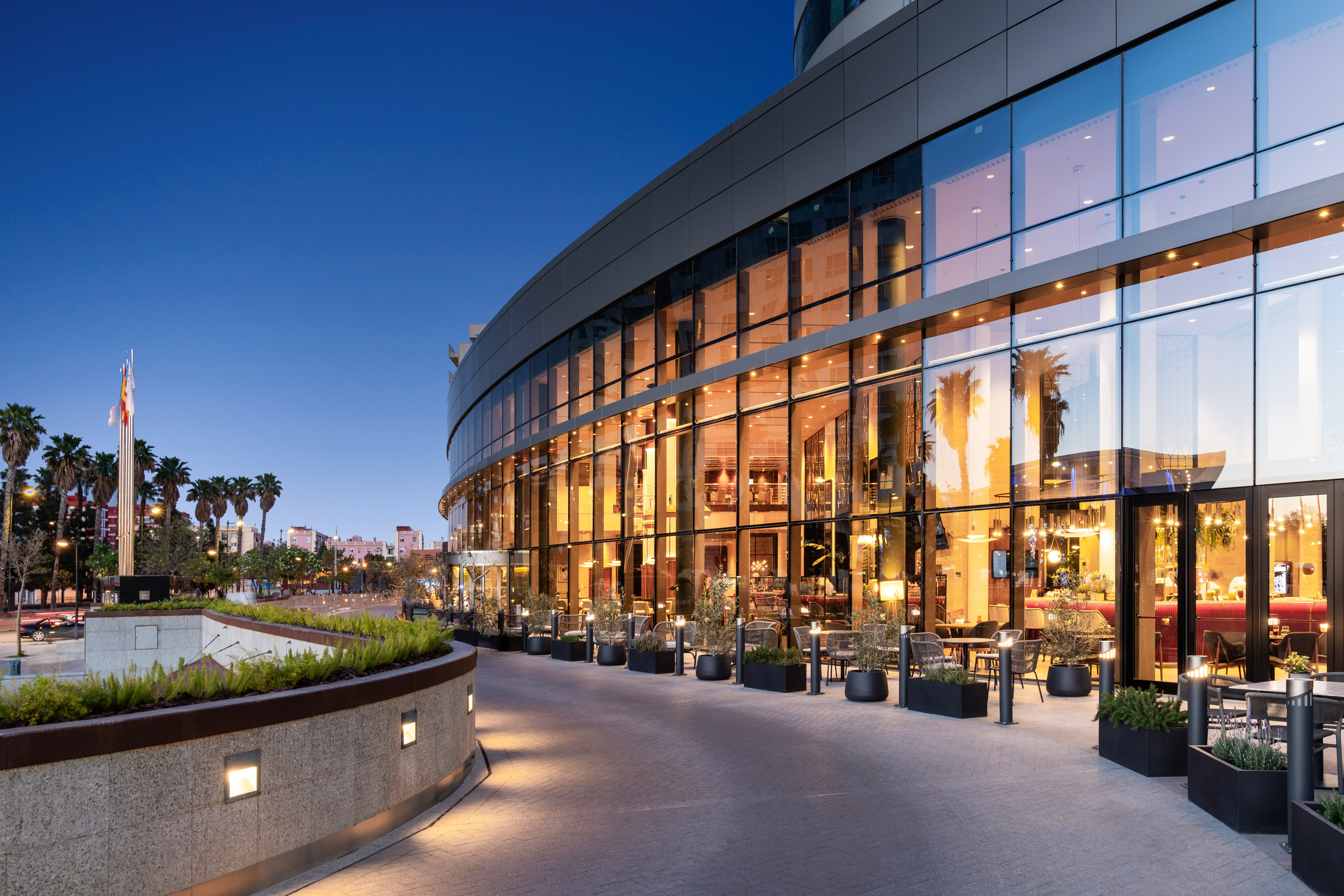 Modern curved glass hotel exterior at twilight with outdoor seating and palm trees.