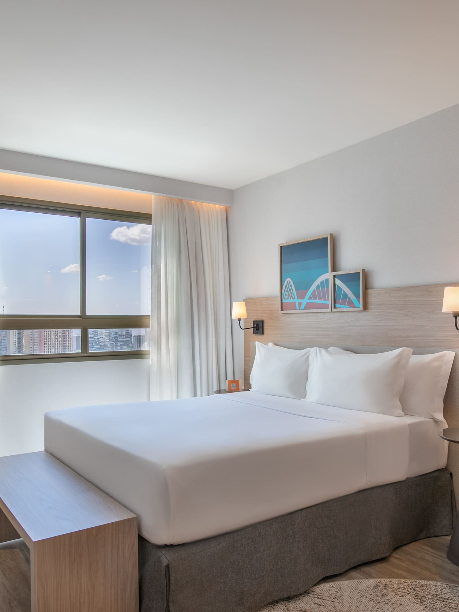 Modern hotel bedroom with white bed, cityscape view, and contemporary furnishings.