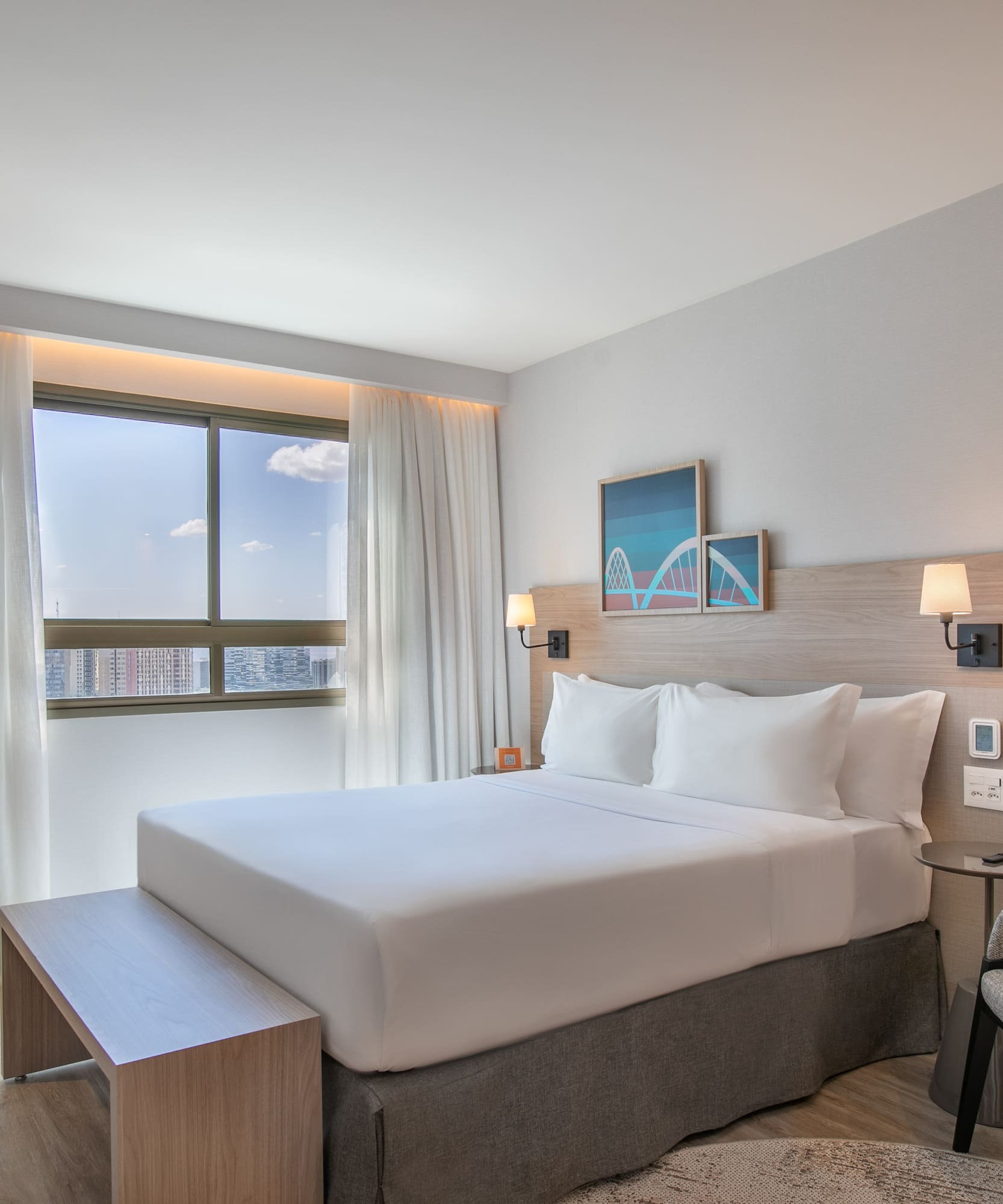 Modern hotel bedroom with white bed, cityscape view, and contemporary furnishings.