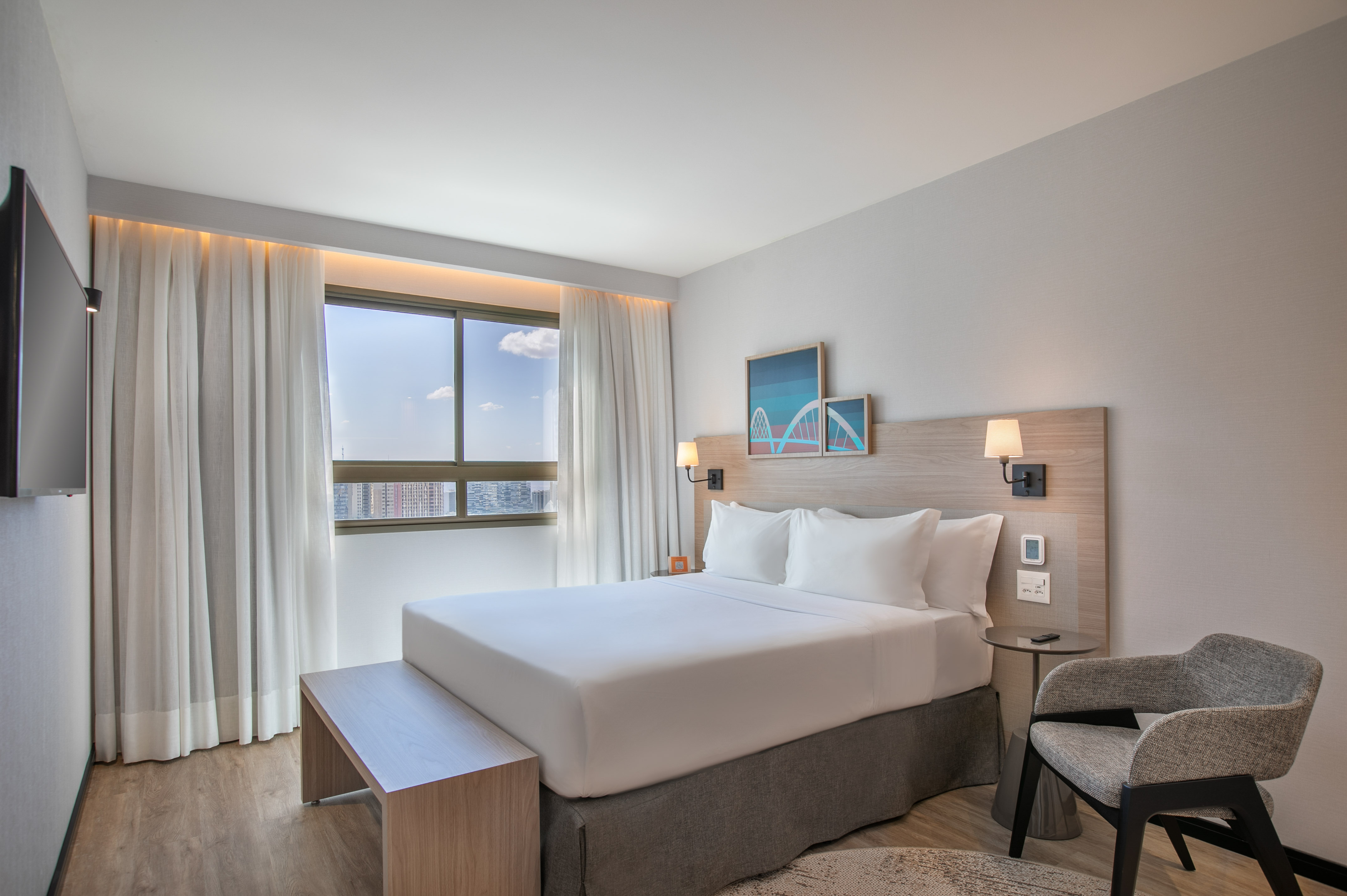 Modern hotel bedroom with white bed, cityscape view, and contemporary furnishings.