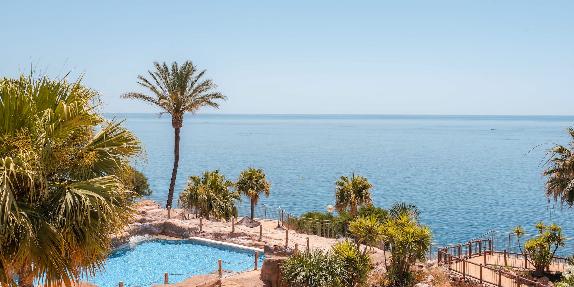 Tropical resort pool with ocean view, surrounded by palm trees and rocks.