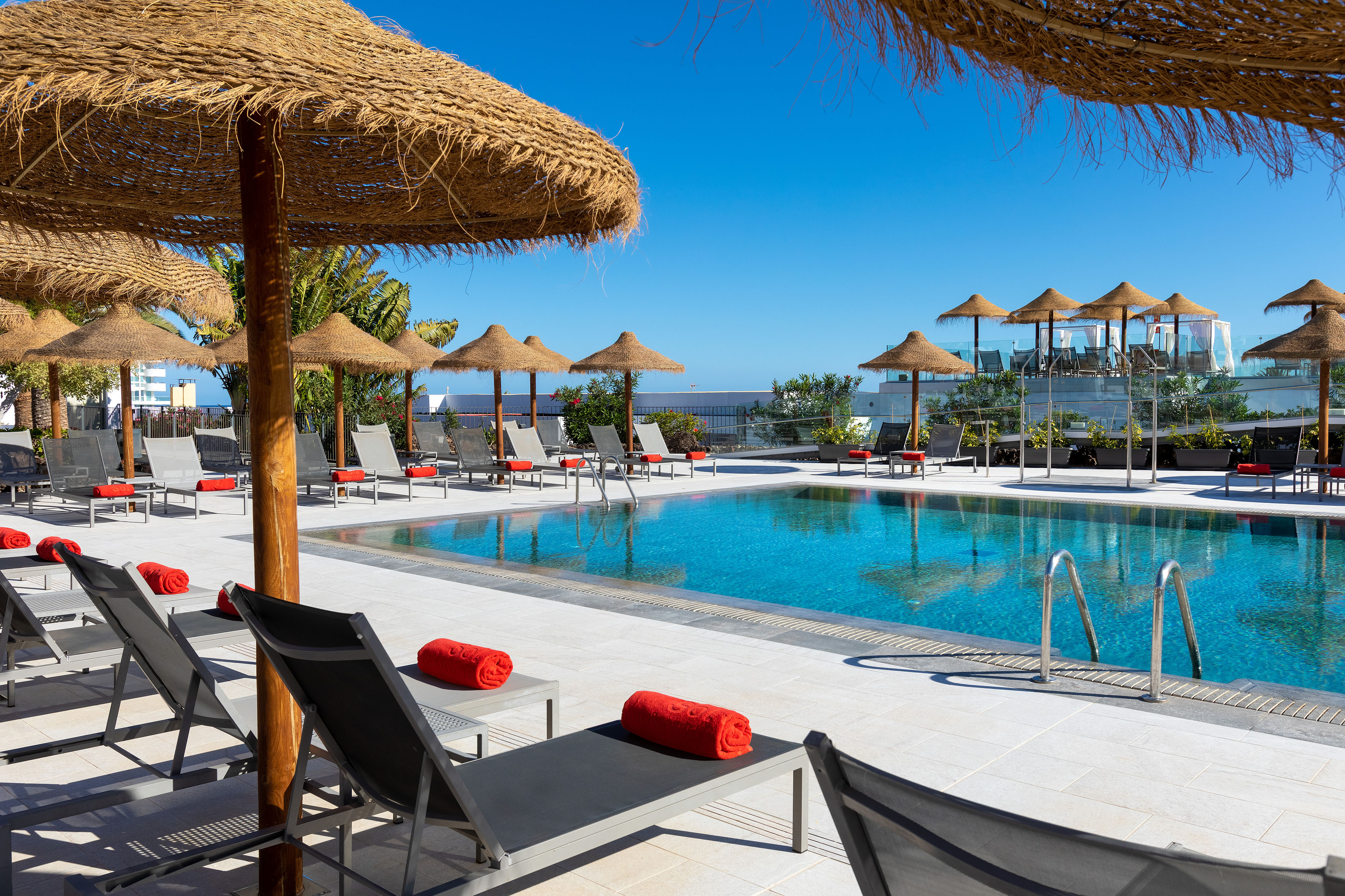 Sunny resort swimming pool with gray loungers, red towels, and thatched umbrellas.
