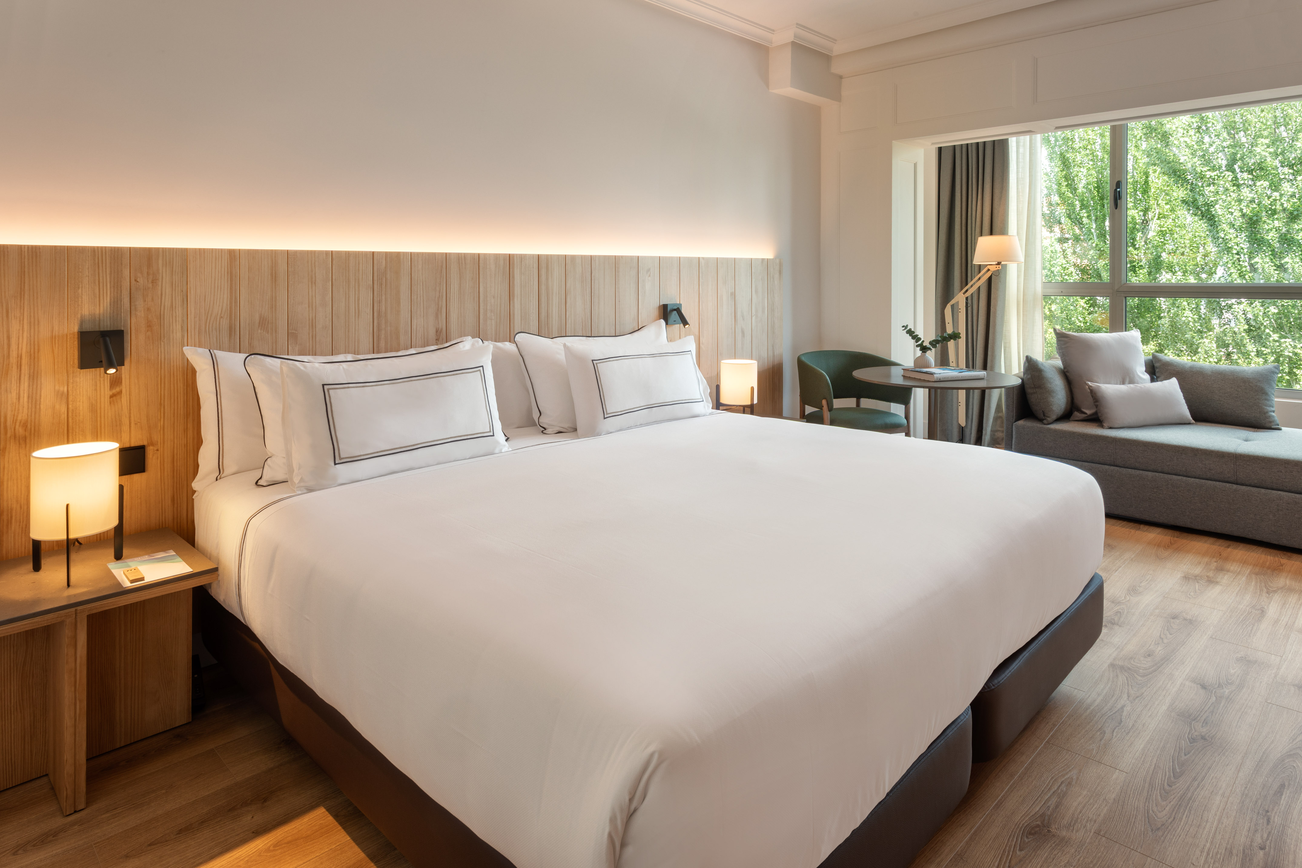 Modern hotel room with king bed, wooden headboard, window, and comfortable seating.