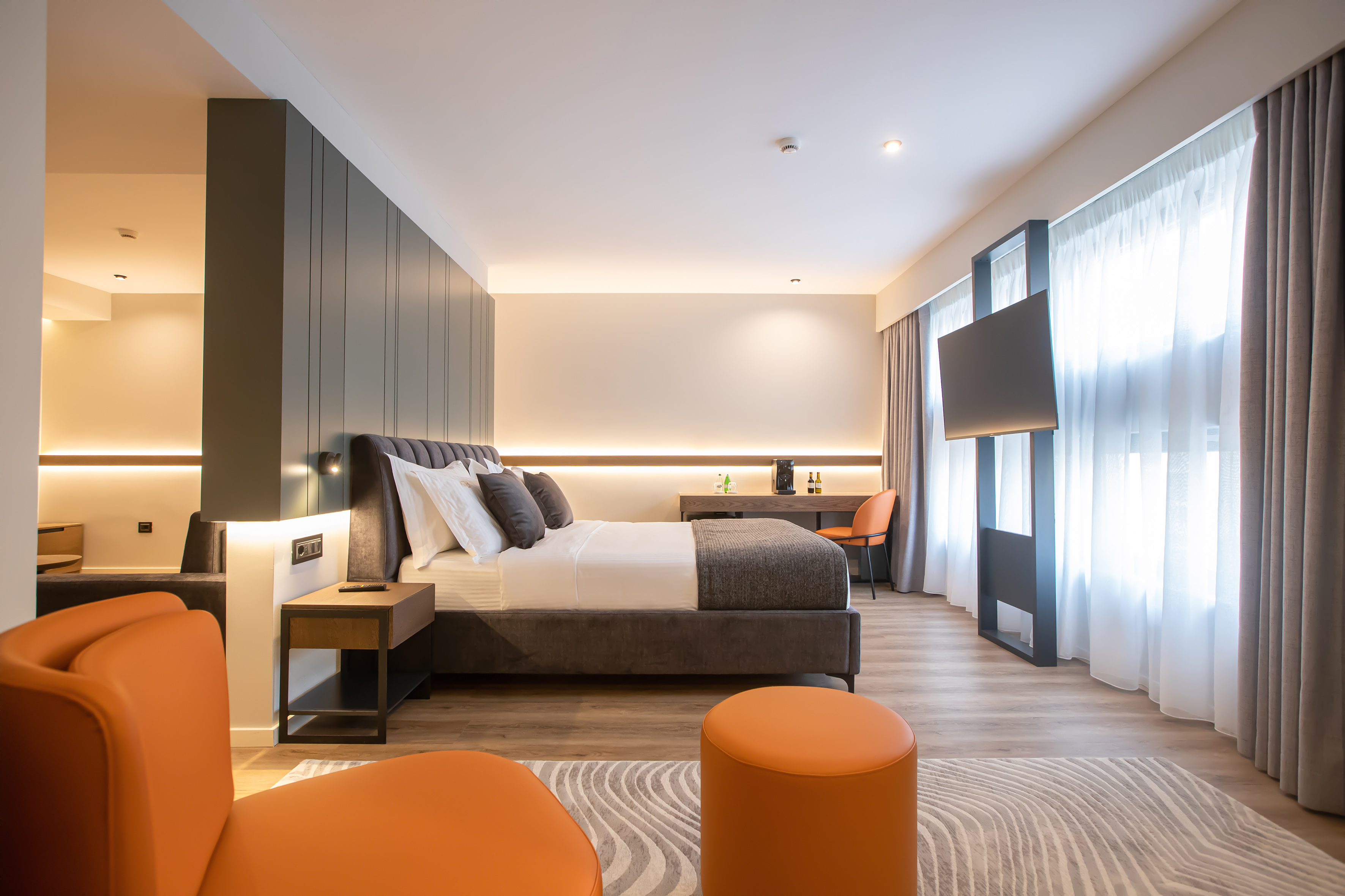 Modern hotel bedroom with large bed, orange chair, desk, and wall-mounted TV.