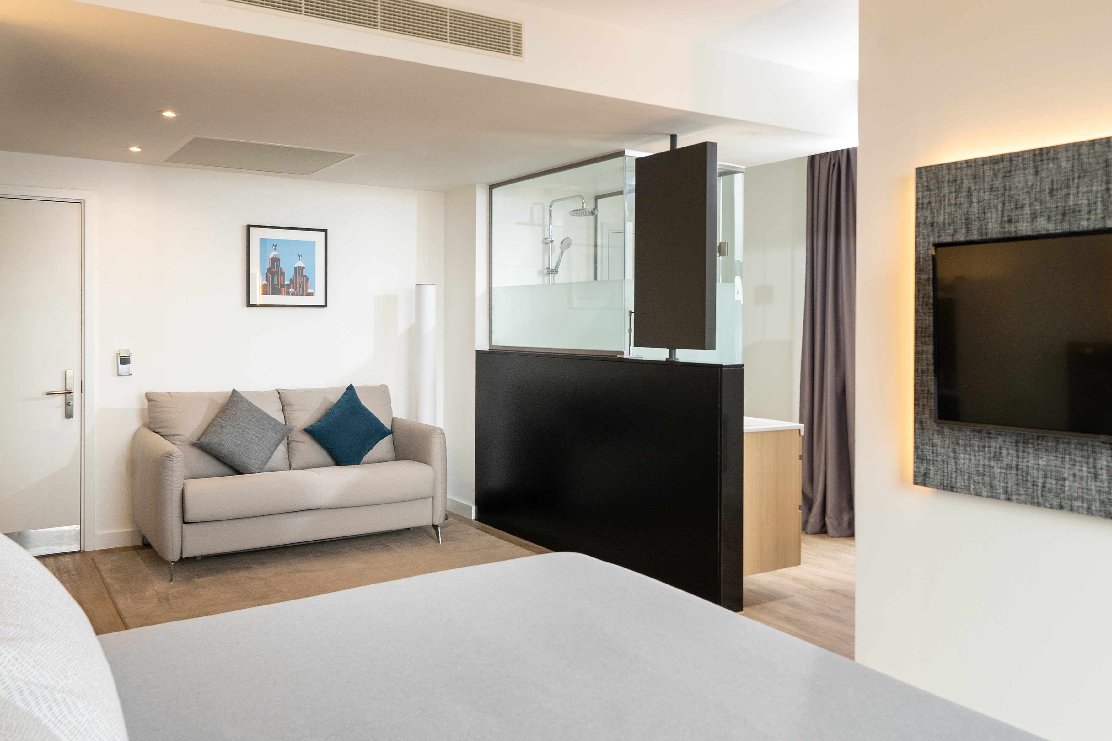 Modern hotel suite interior with sofa, glass shower, and backlit TV.