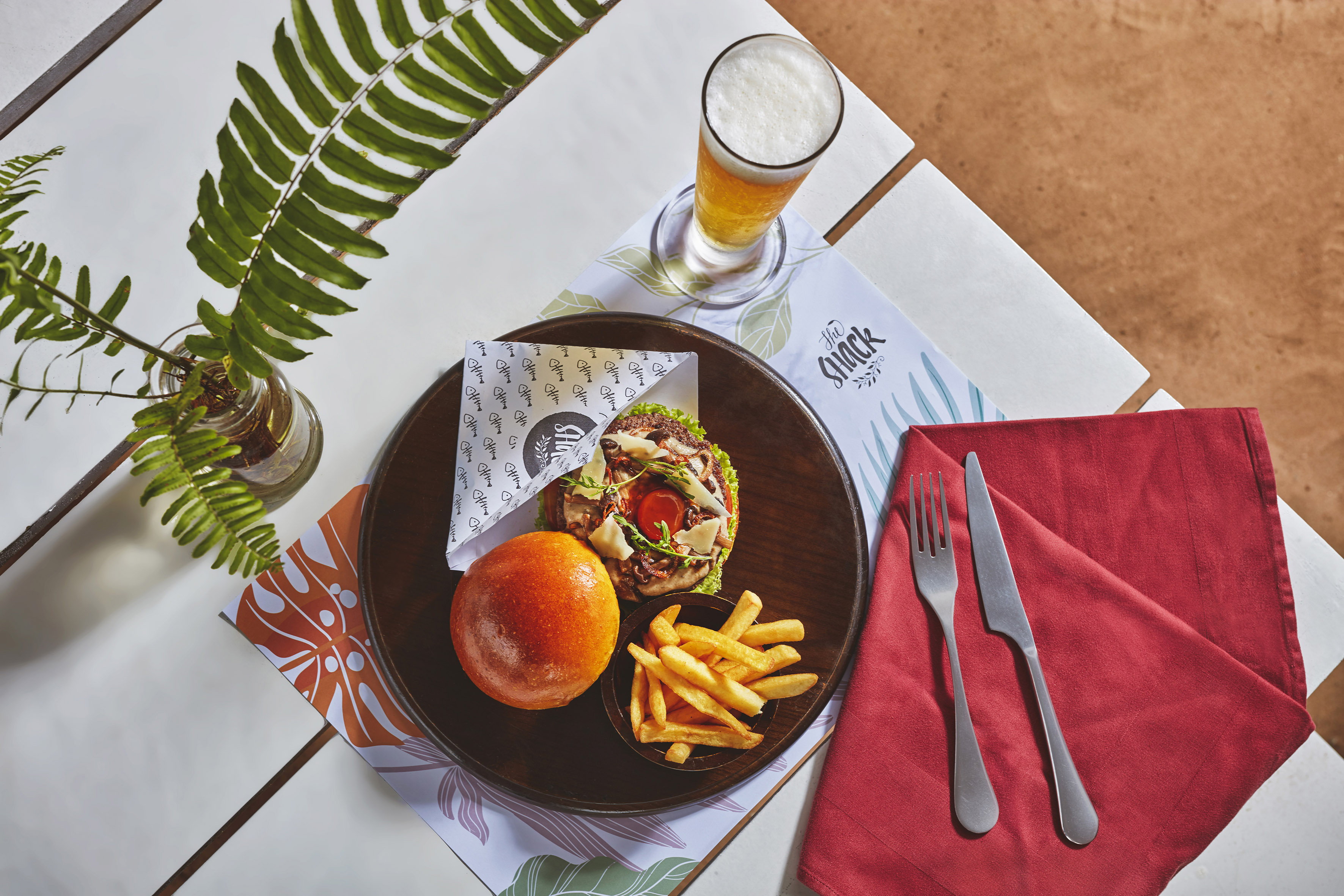 Gourmet burger, crispy fries, and beer on a wooden tray with fern decor.
