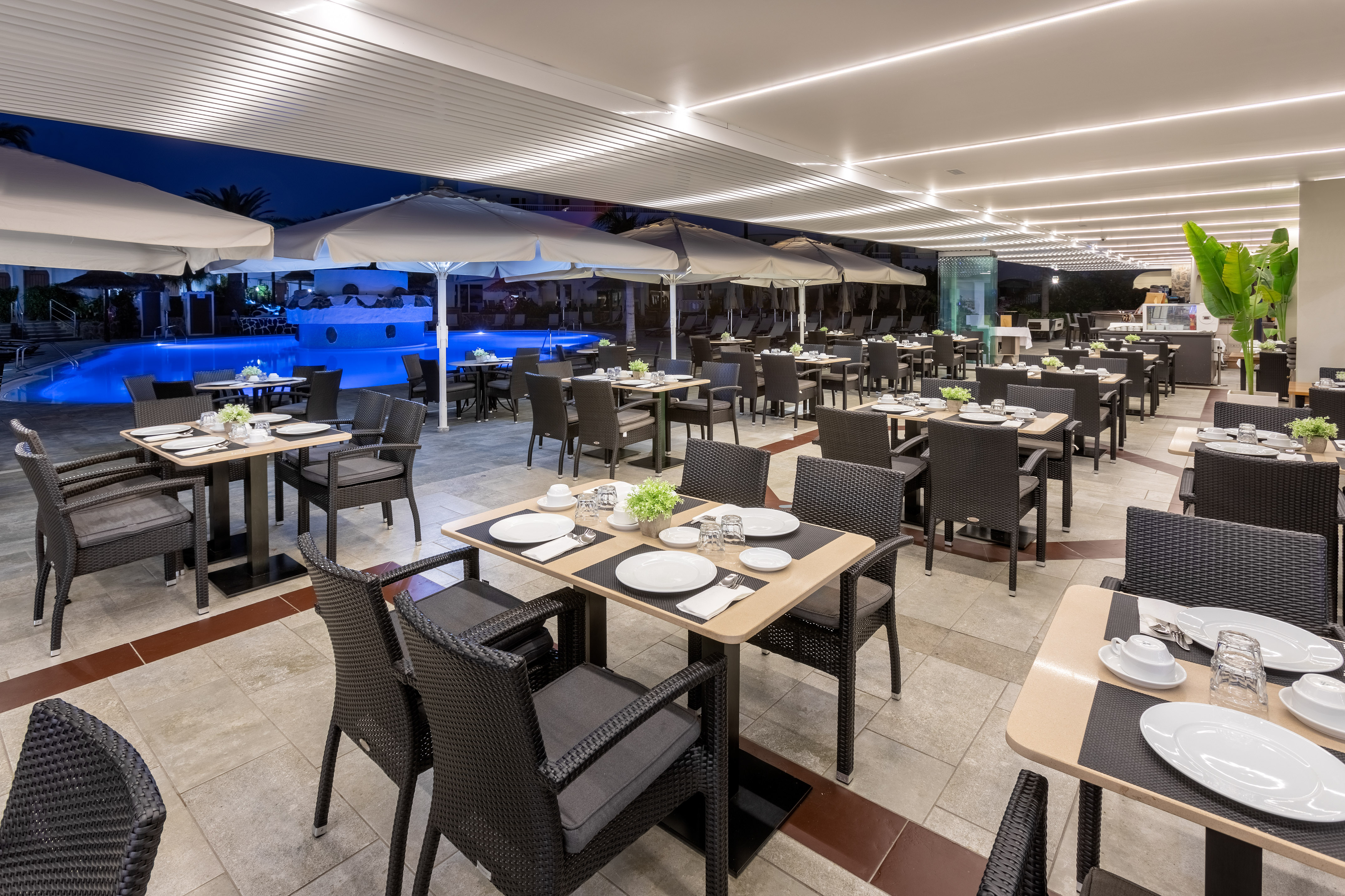 Outdoor restaurant by illuminated pool at night, modern tables & chairs.