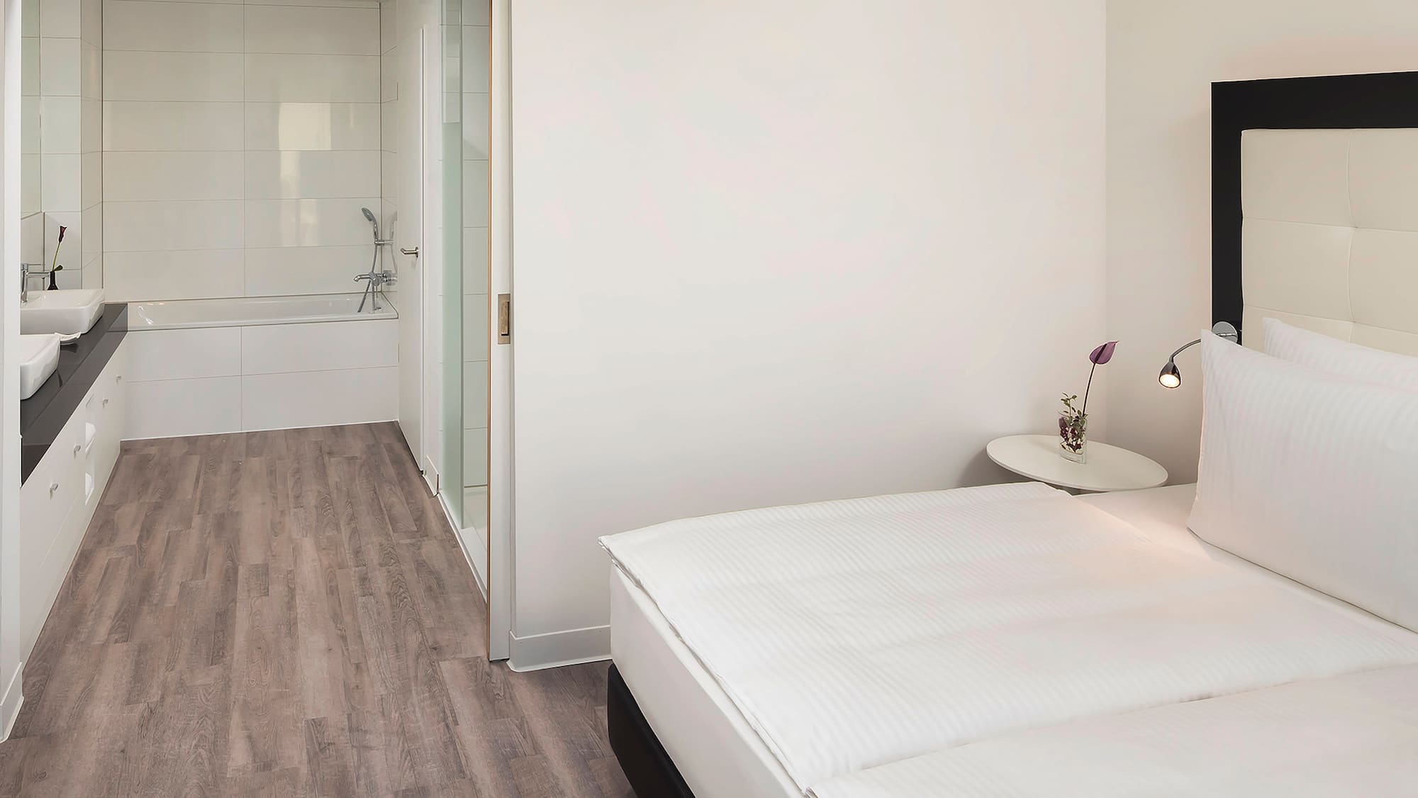 Modern hotel bedroom with white bed, nightstand, and bathroom with bathtub.