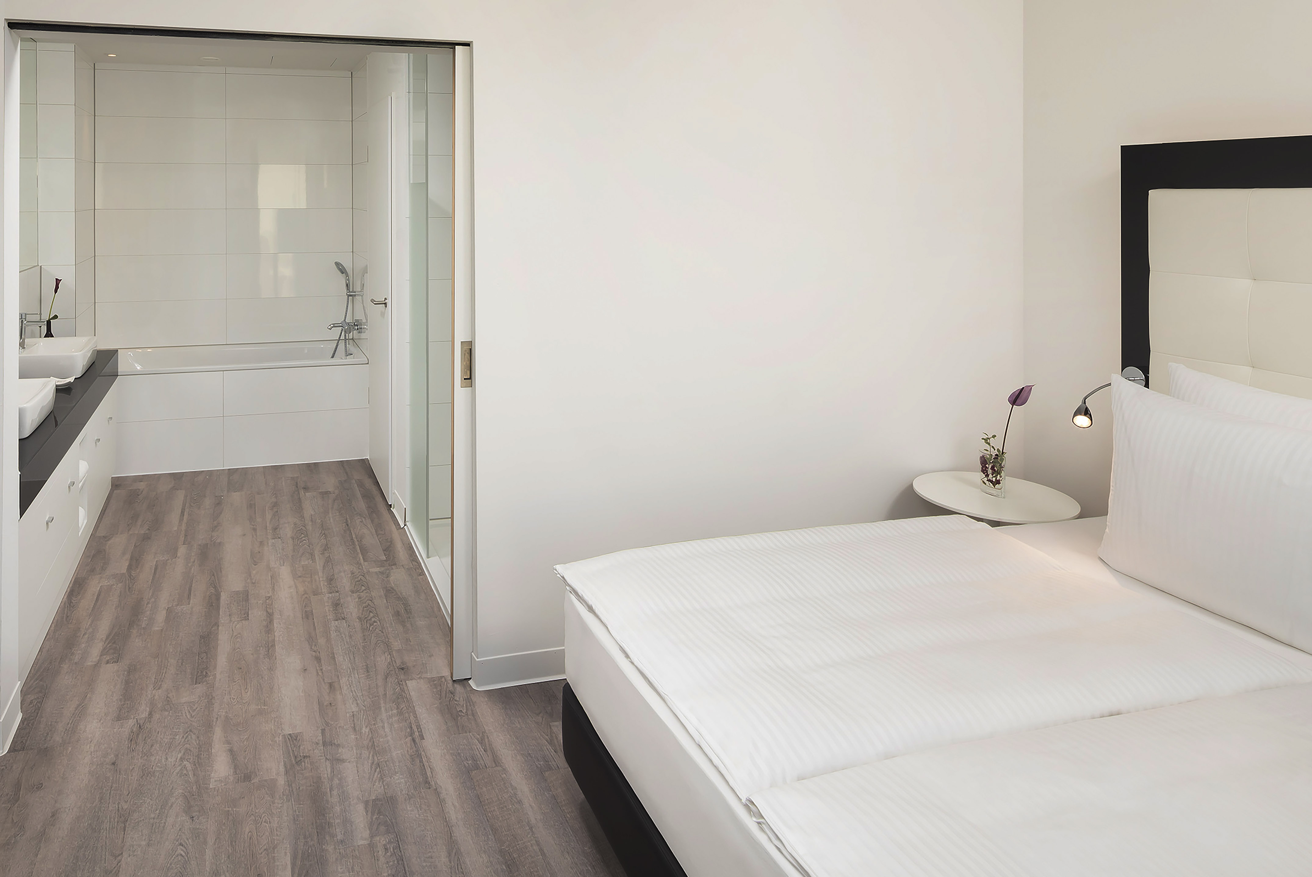 Modern hotel bedroom with white bed, nightstand, and bathroom with bathtub.