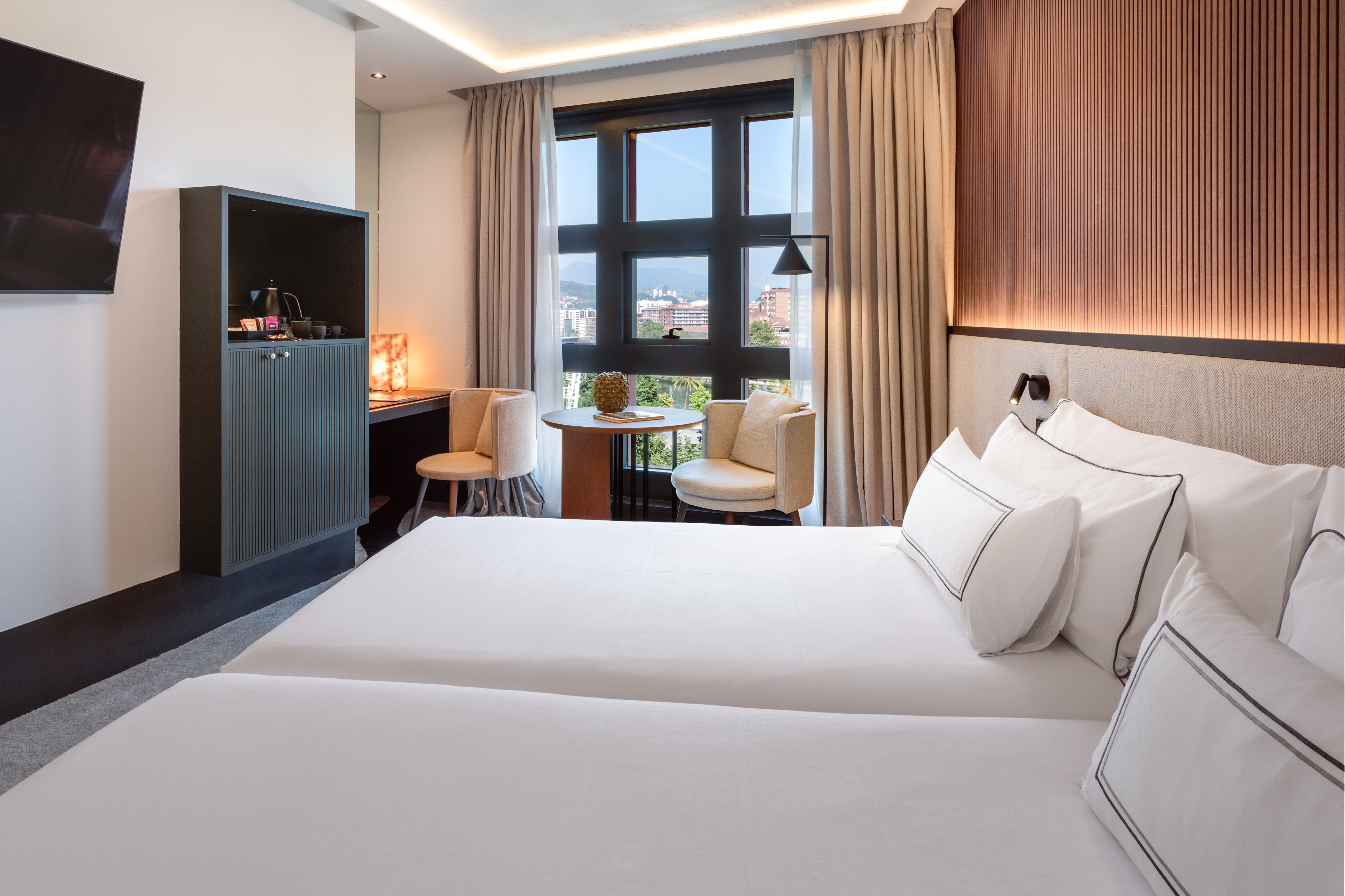 Modern hotel room with twin beds, city view, and coffee station.