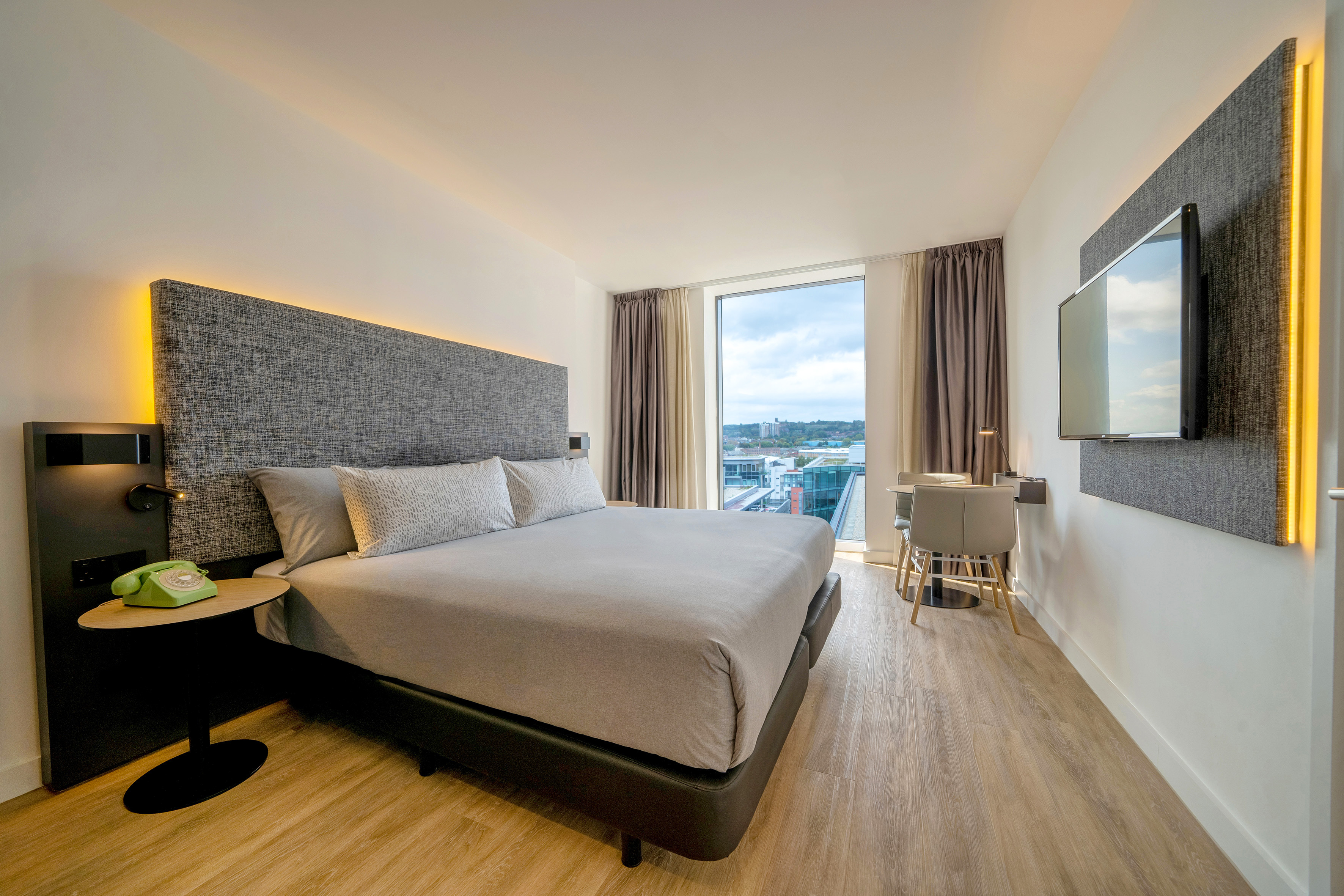 Modern hotel room with king bed, backlit headboard, city view, and desk.