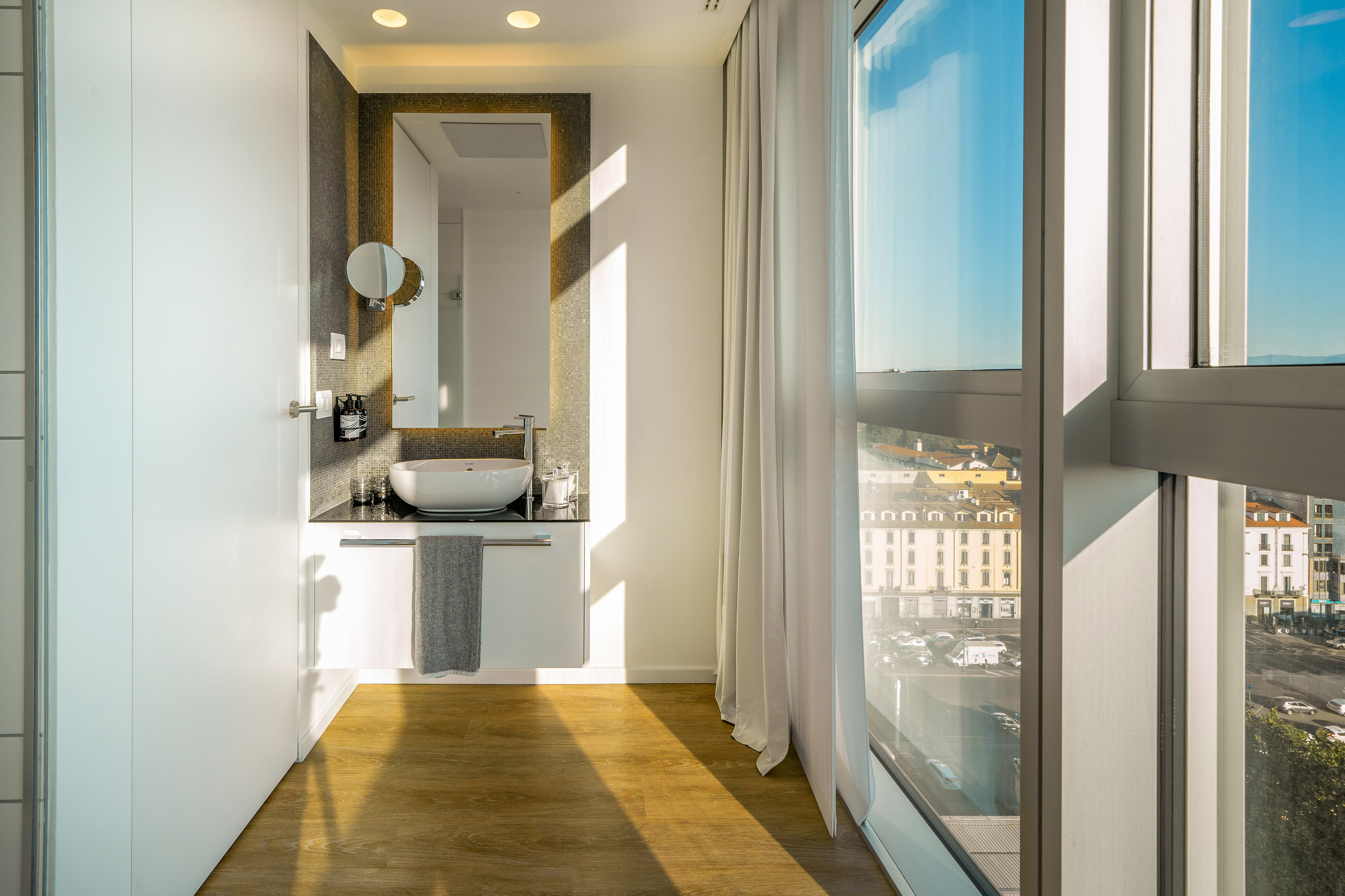 Modern bathroom vanity with vessel sink, large mirror, wood floor, and city view window.