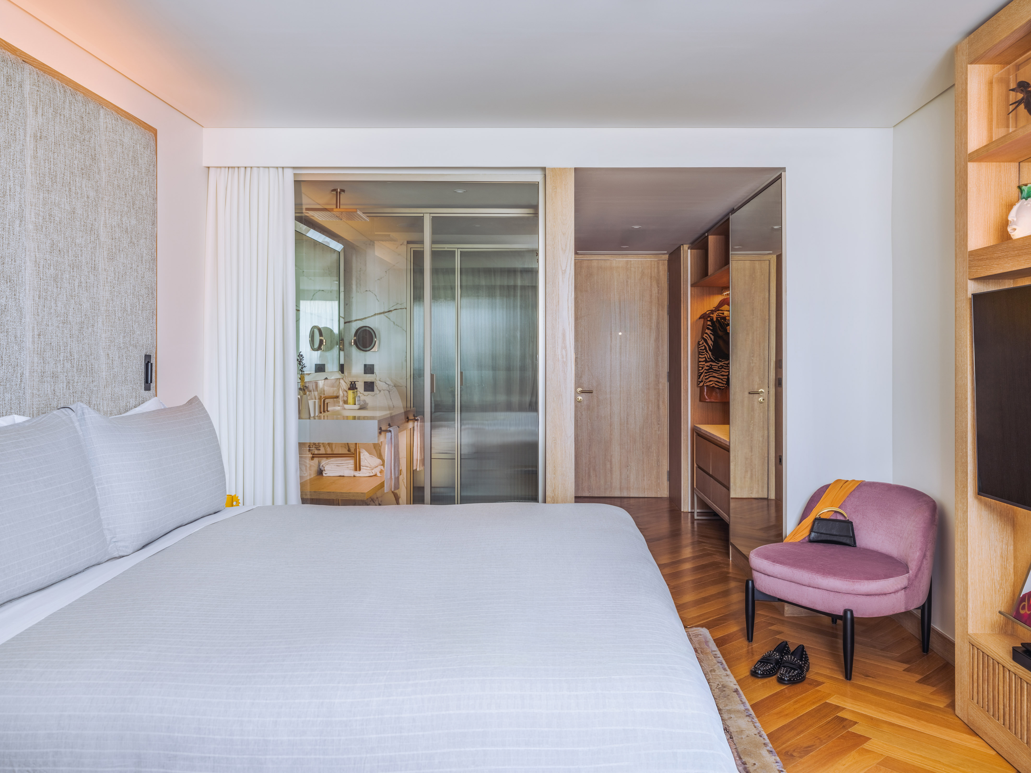 Modern hotel bedroom with ensuite bathroom, walk-in closet, and pink accent chair.
