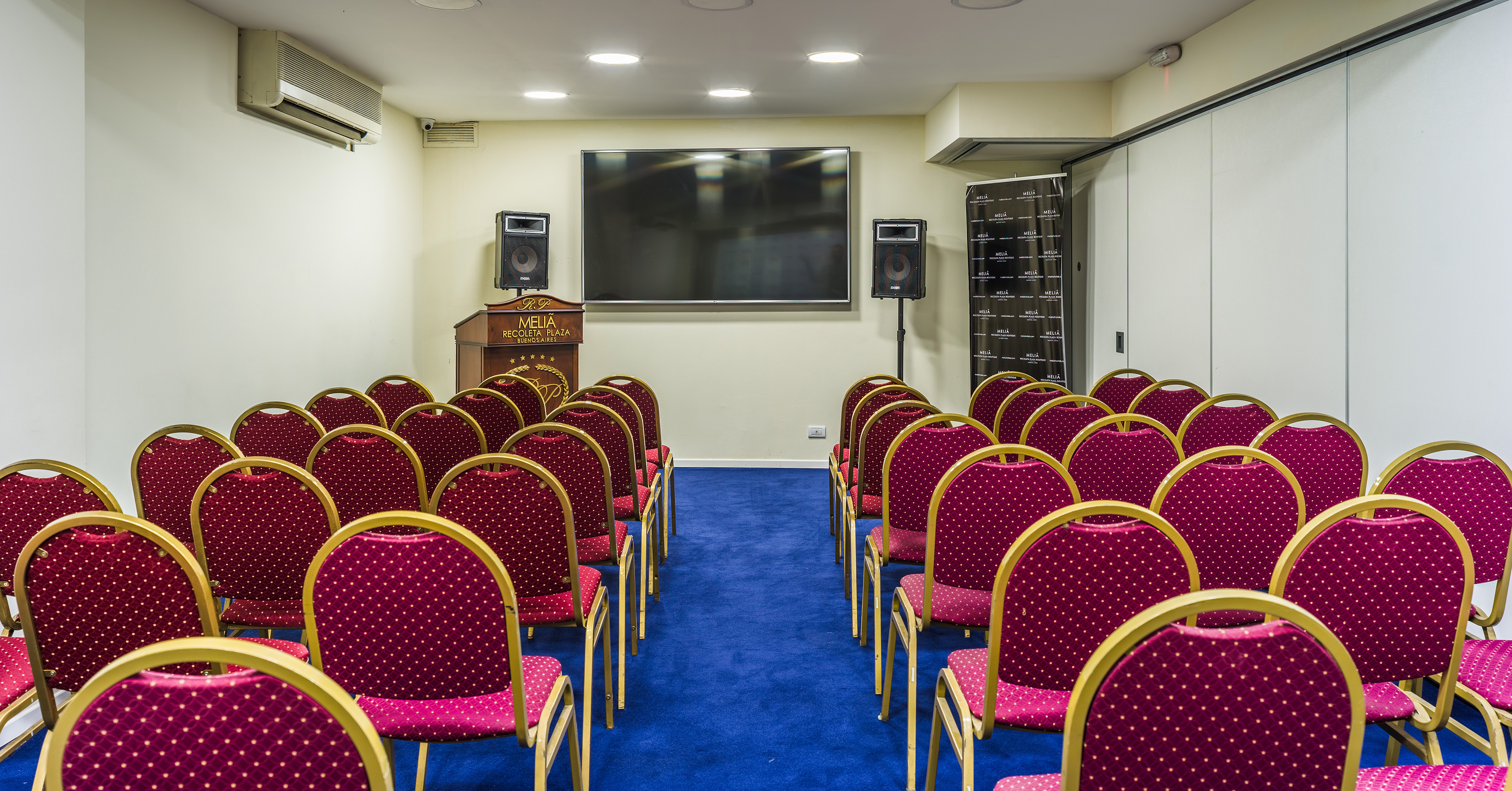 Melia Recoleta Plaza meeting room with red chairs, blue carpet, screen, and podium.