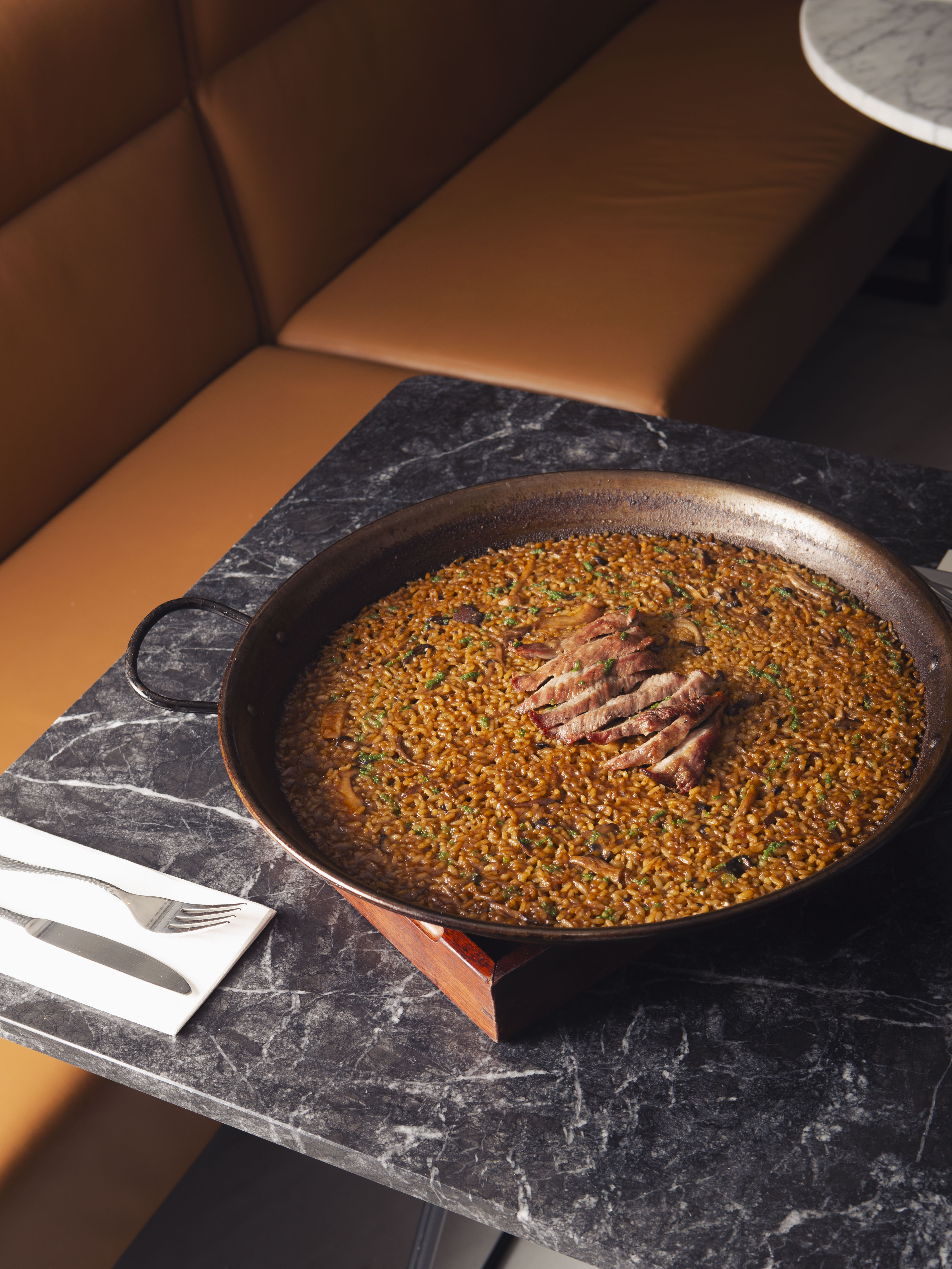 Large paella with sliced meat and green herbs on a dark marble table.