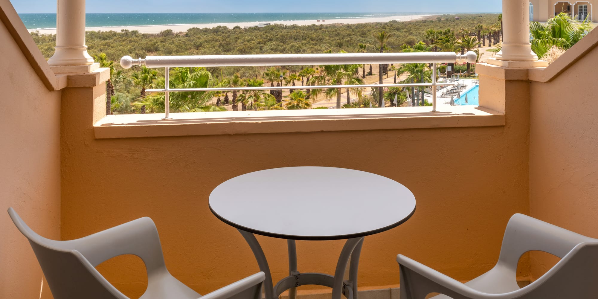 Hotel balcony overlooking ocean beach, palm trees, and swimming pool.