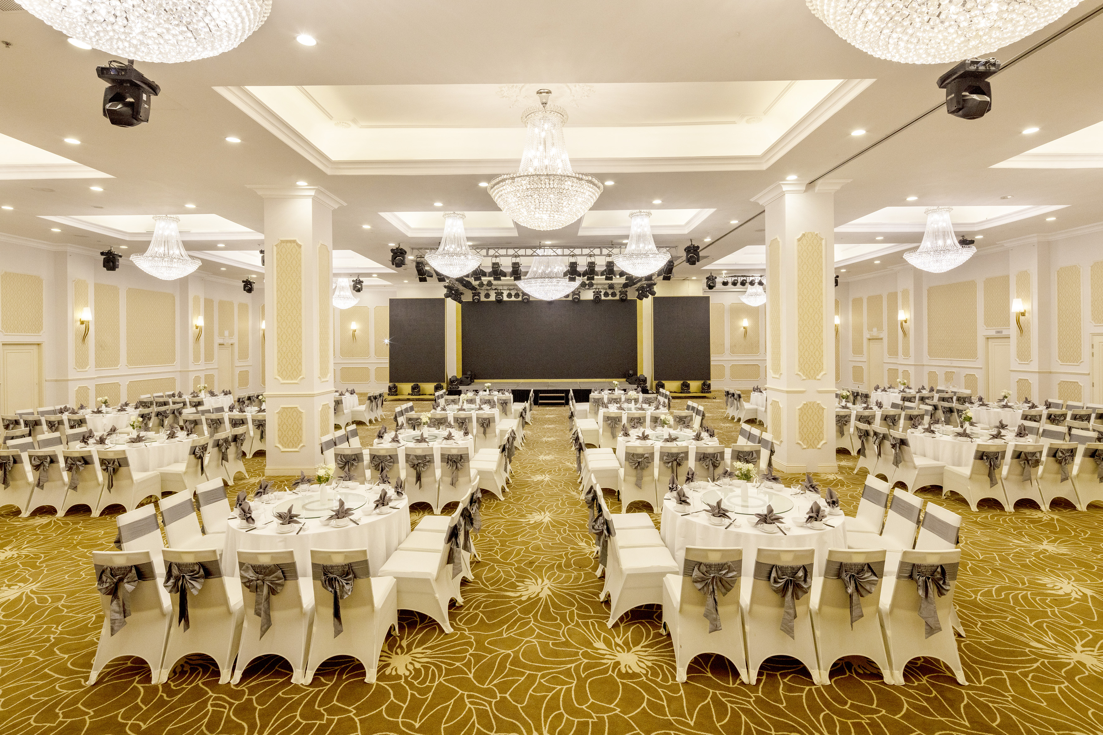 Elegant banquet hall with round dining tables, white chairs, chandeliers, and stage.