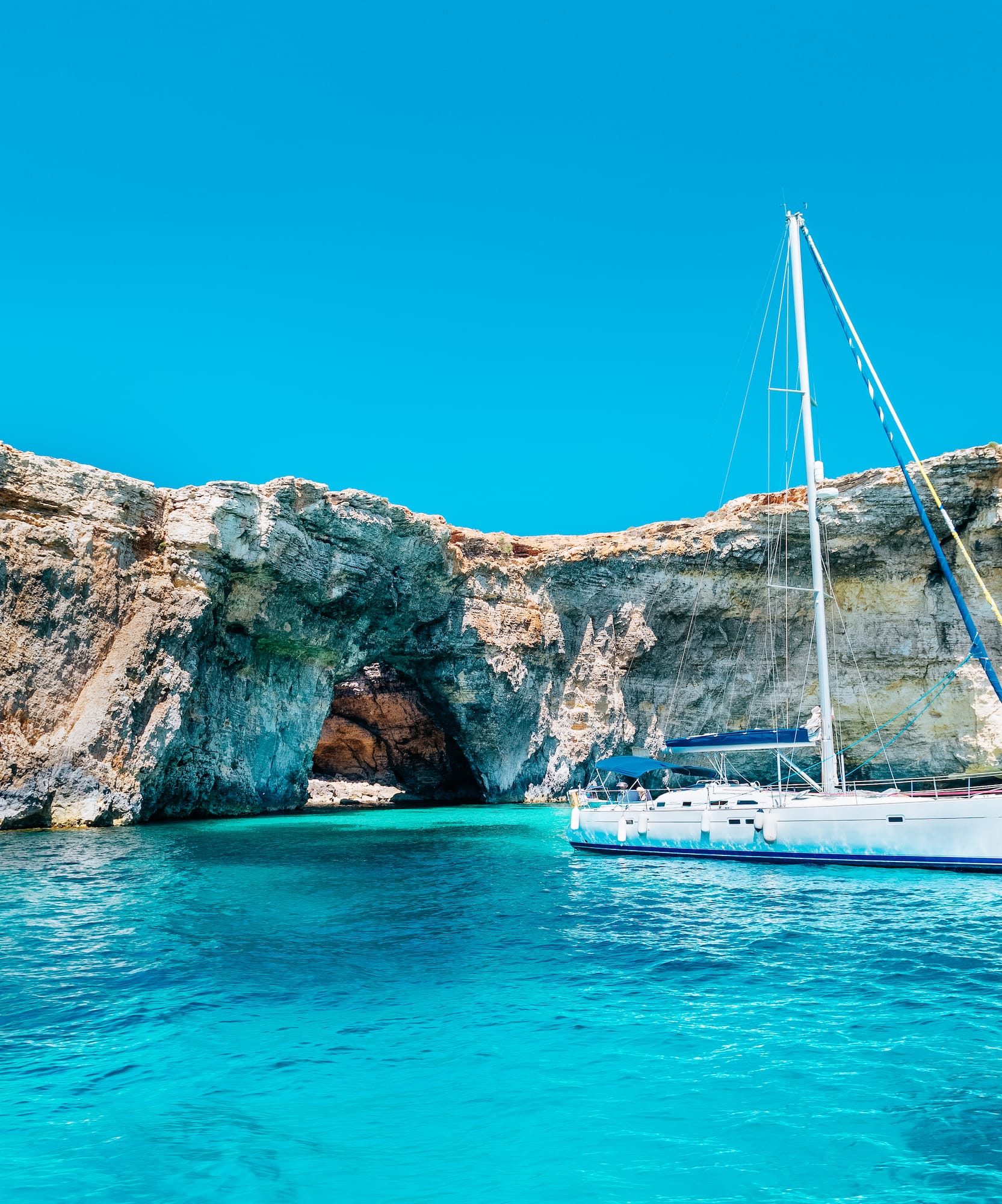 White sailboat in turquoise water near rocky cliffs and sea cave.