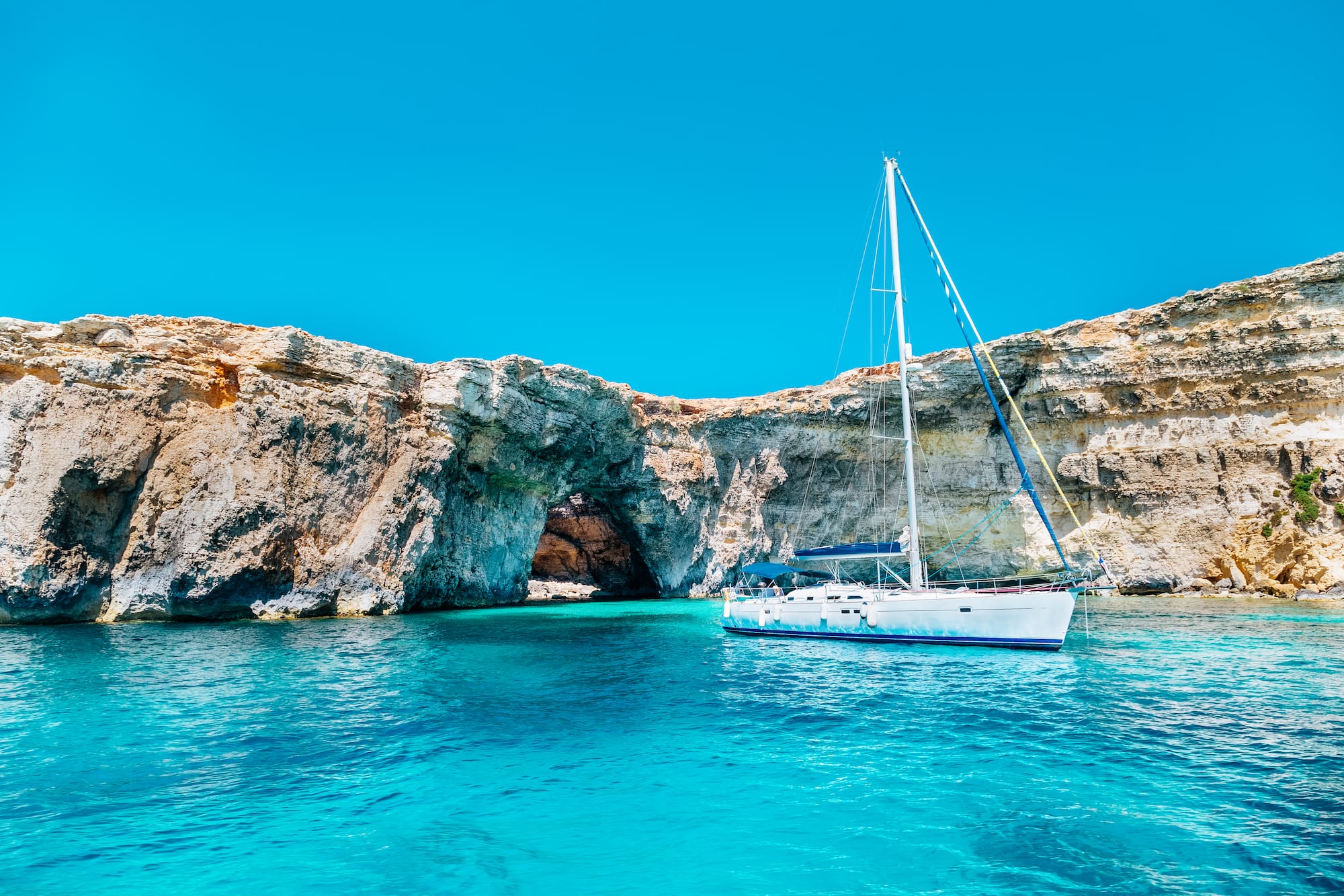 Sailboat in clear turquoise water by rocky cliffs and natural arch.