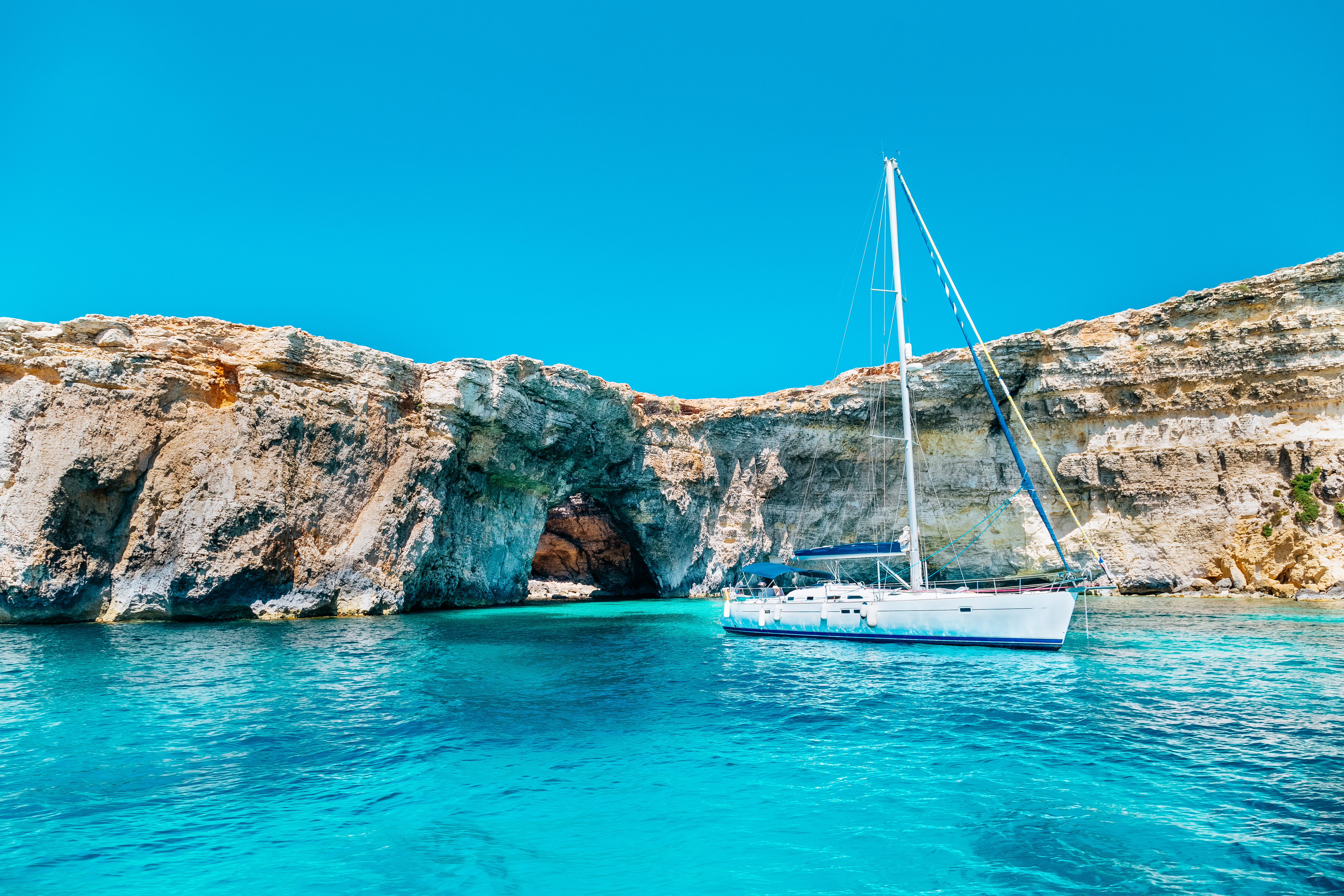 White sailboat in turquoise water near rocky cliffs and sea cave.