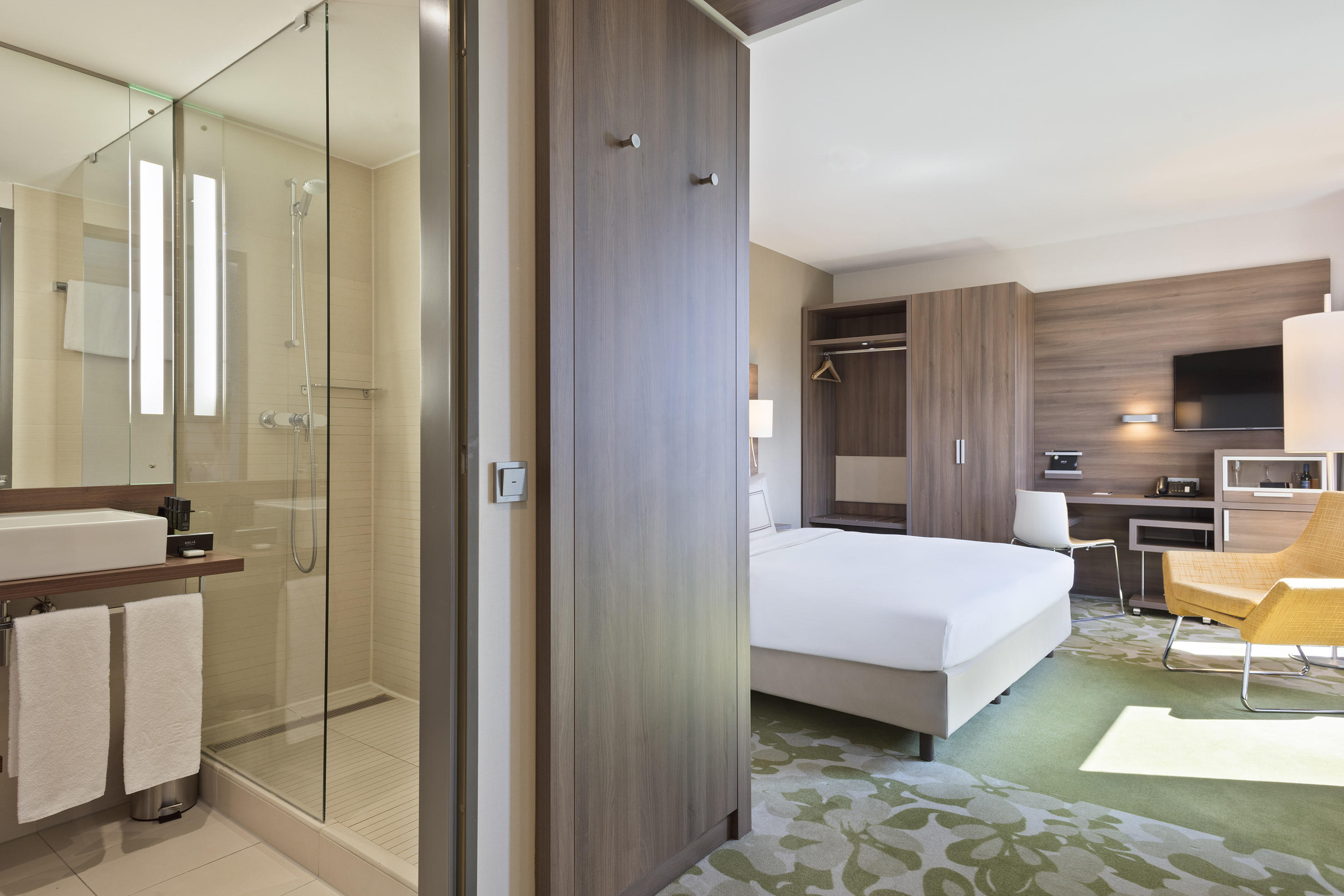Modern hotel room with bed, desk, and bathroom with glass shower.