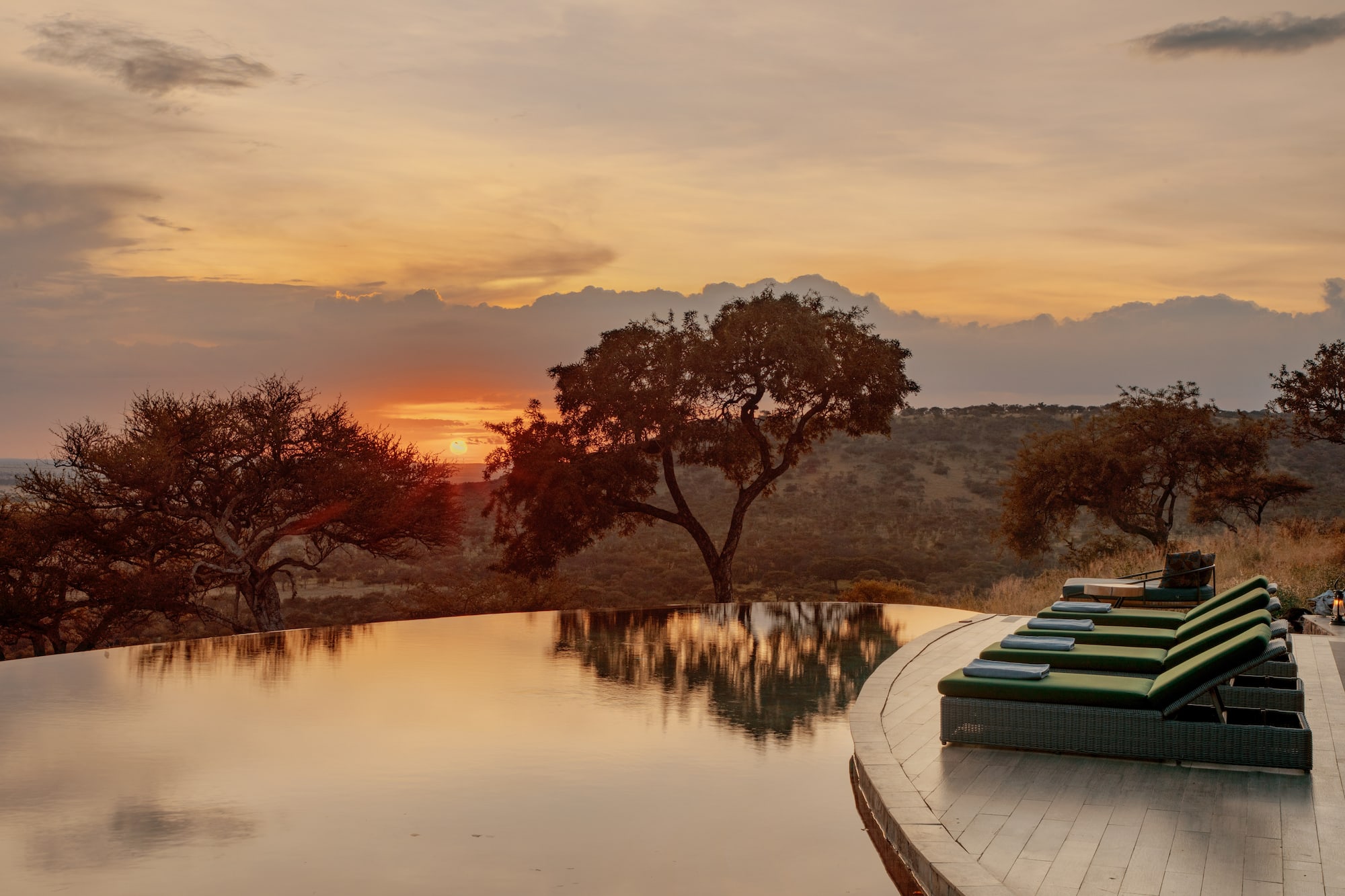 African savanna sunset reflecting in infinity pool with resort lounge chairs.