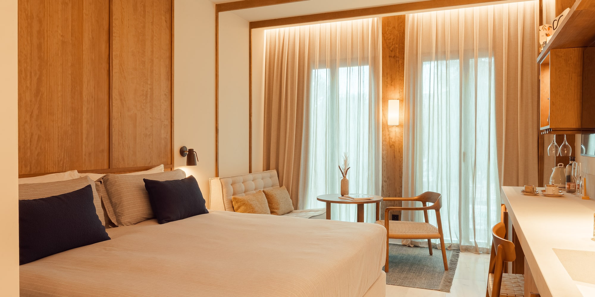 Modern hotel bedroom with large bed, wooden walls, sitting area, and kitchenette.