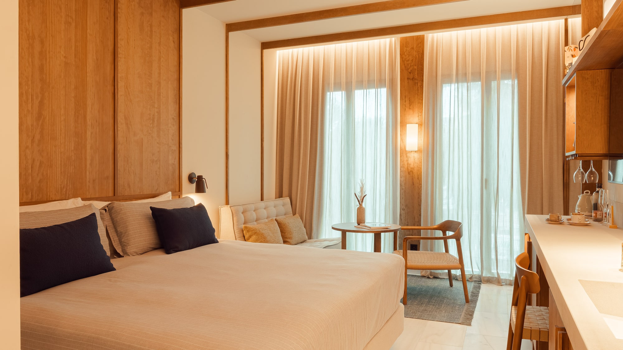 Modern hotel bedroom with large bed, wooden walls, sitting area, and kitchenette.