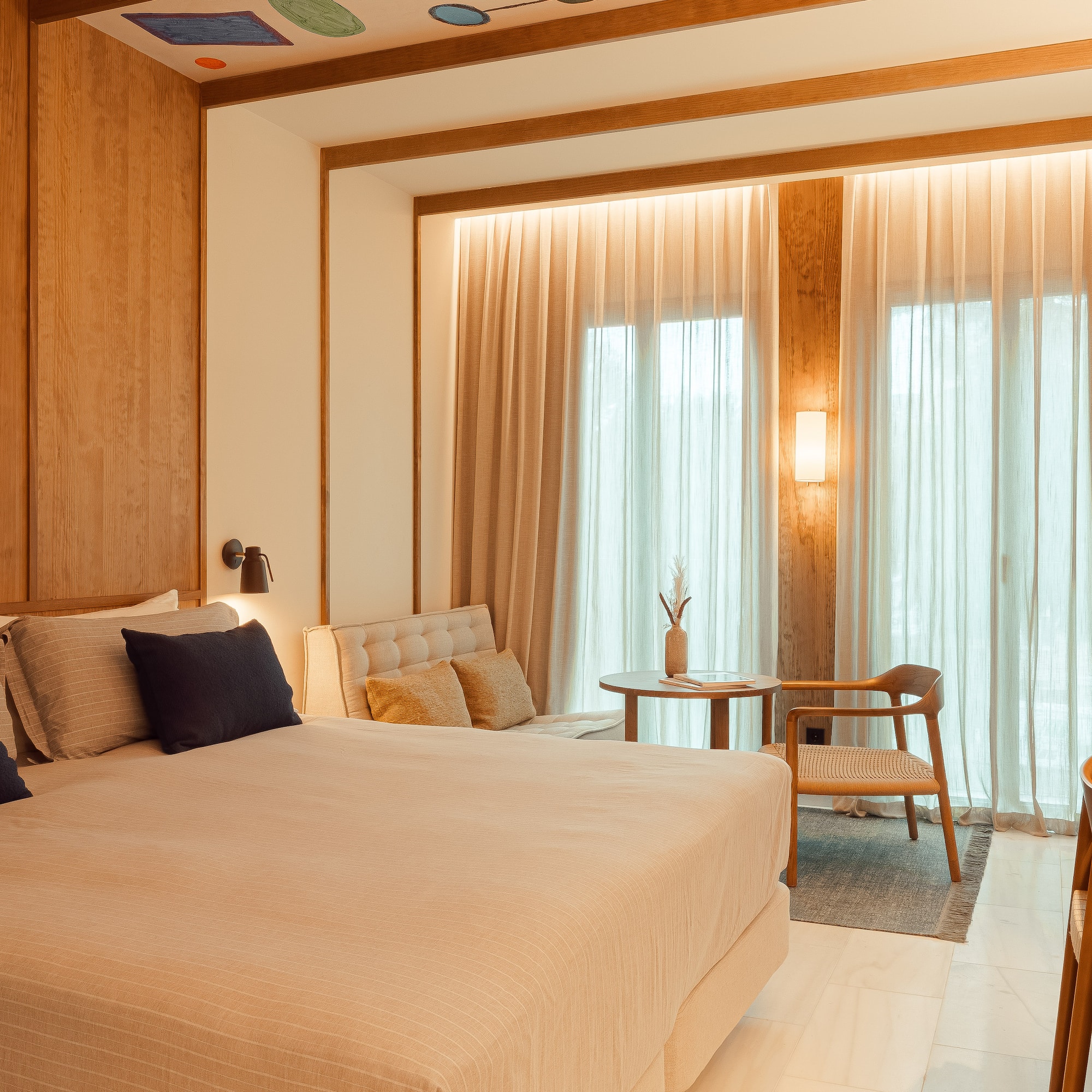 Modern hotel bedroom with large bed, wooden walls, sitting area, and kitchenette.