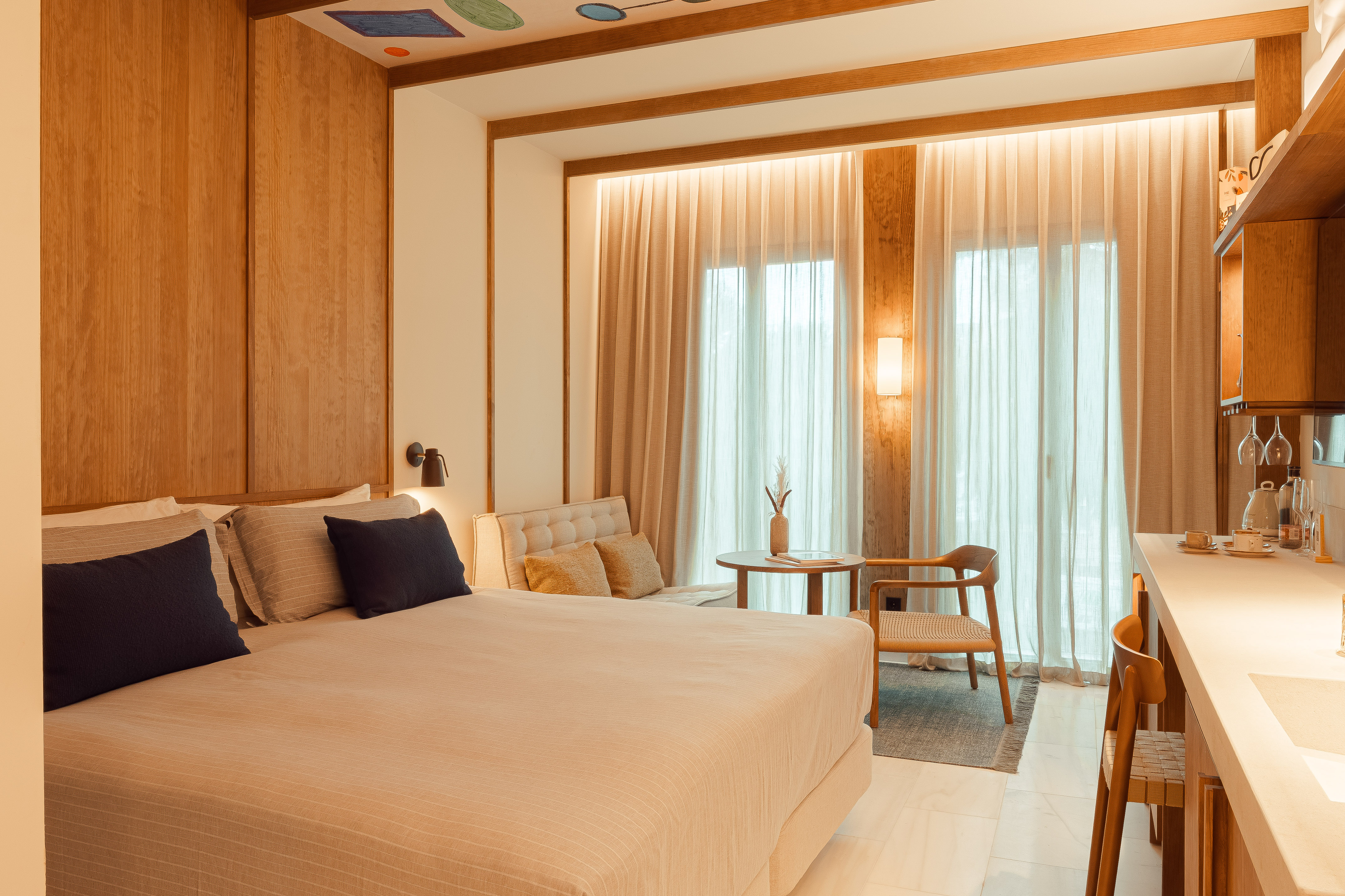 Modern hotel bedroom with large bed, wooden walls, sitting area, and kitchenette.