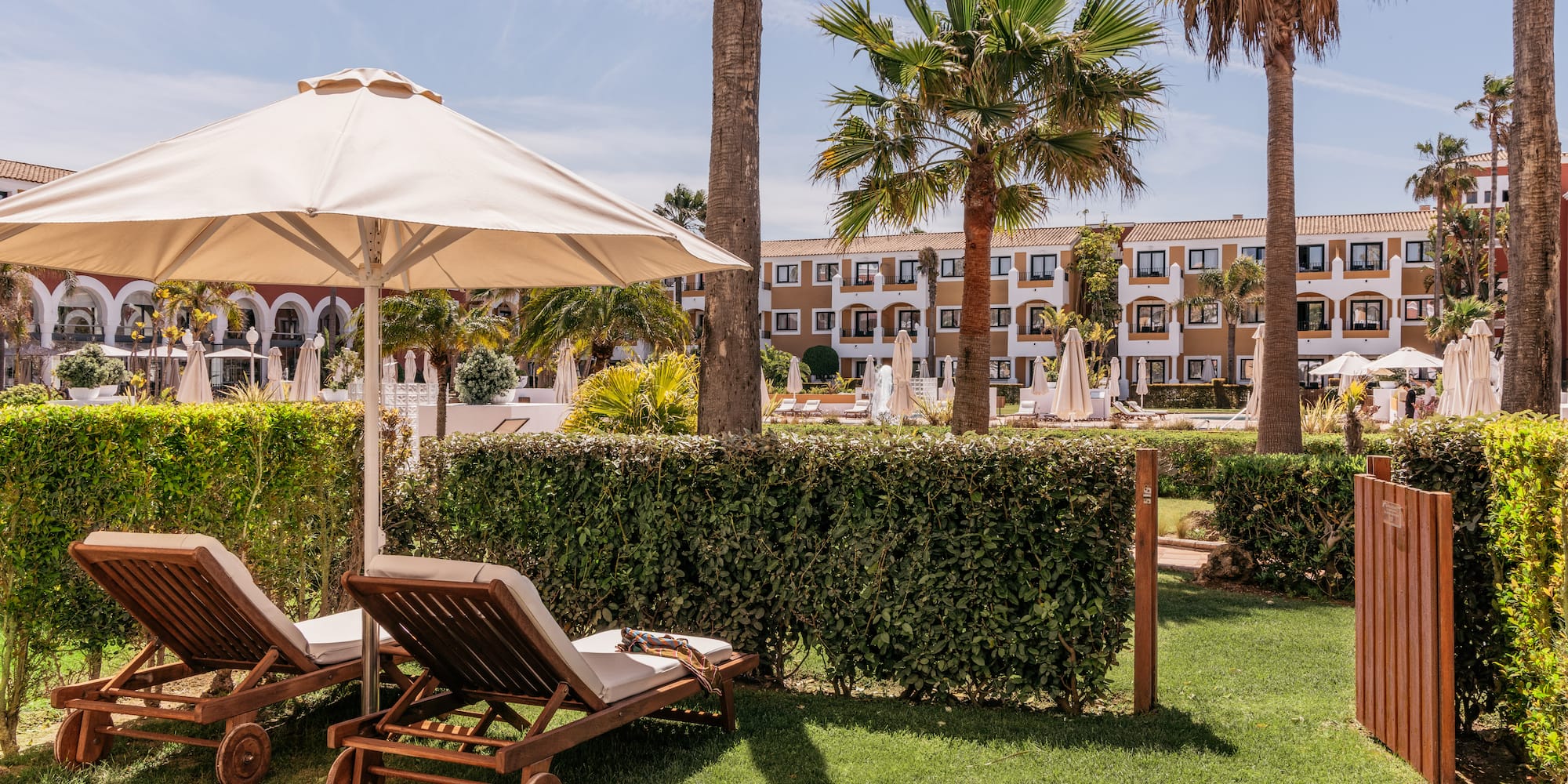 Sunny hotel resort garden, lounge chairs under umbrella, palm trees.