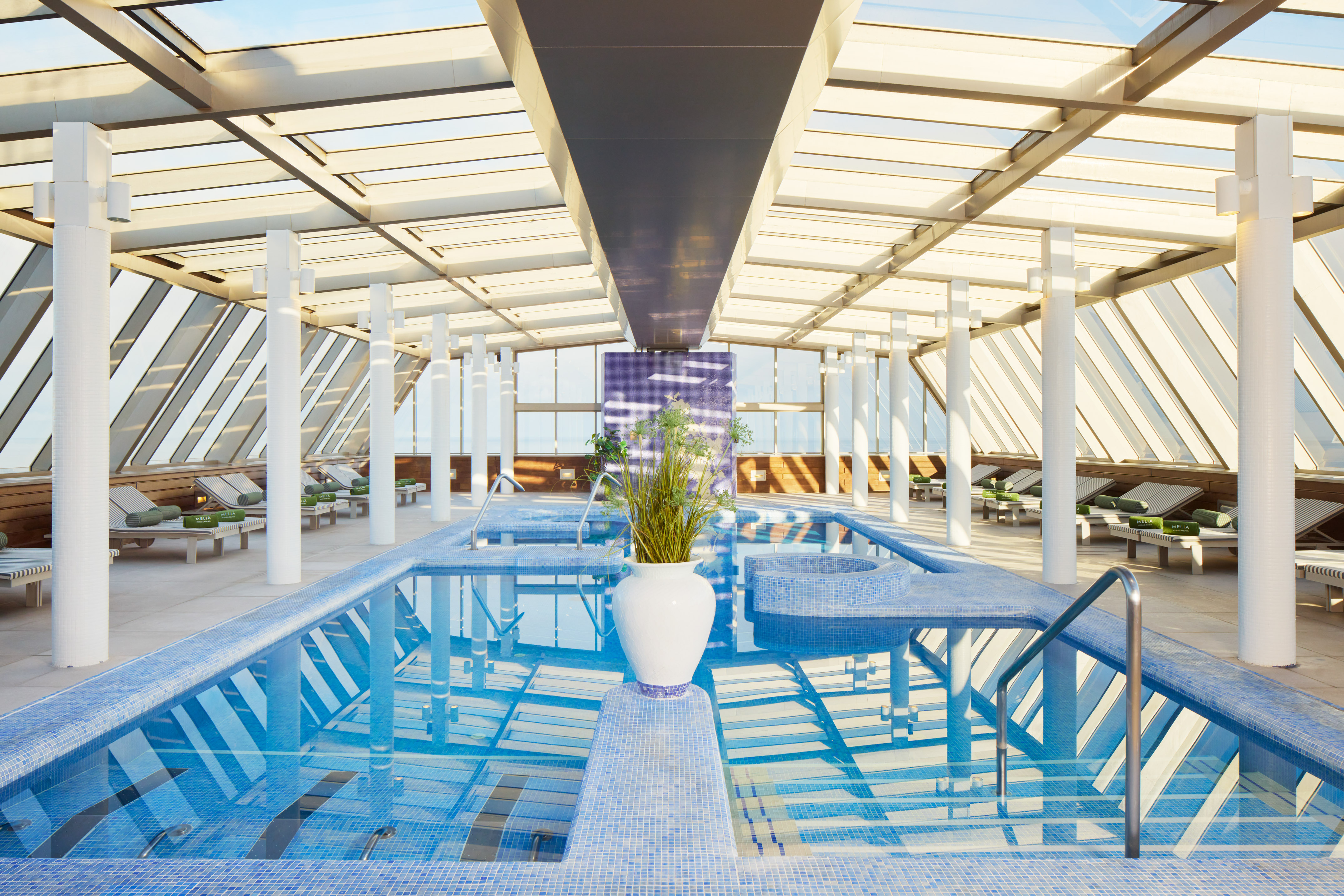 Bright indoor pool with glass roof, blue mosaic tiles, and lounge chairs.