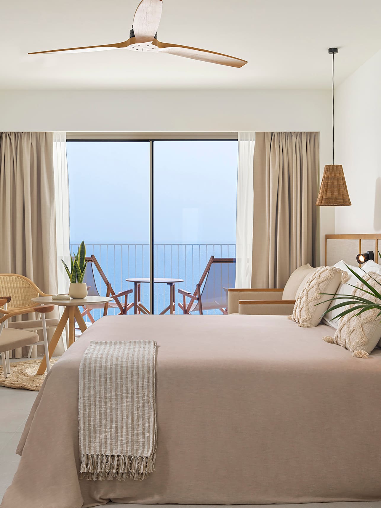 Bright bedroom with ocean view, balcony, neutral decor, and wooden ceiling fan.