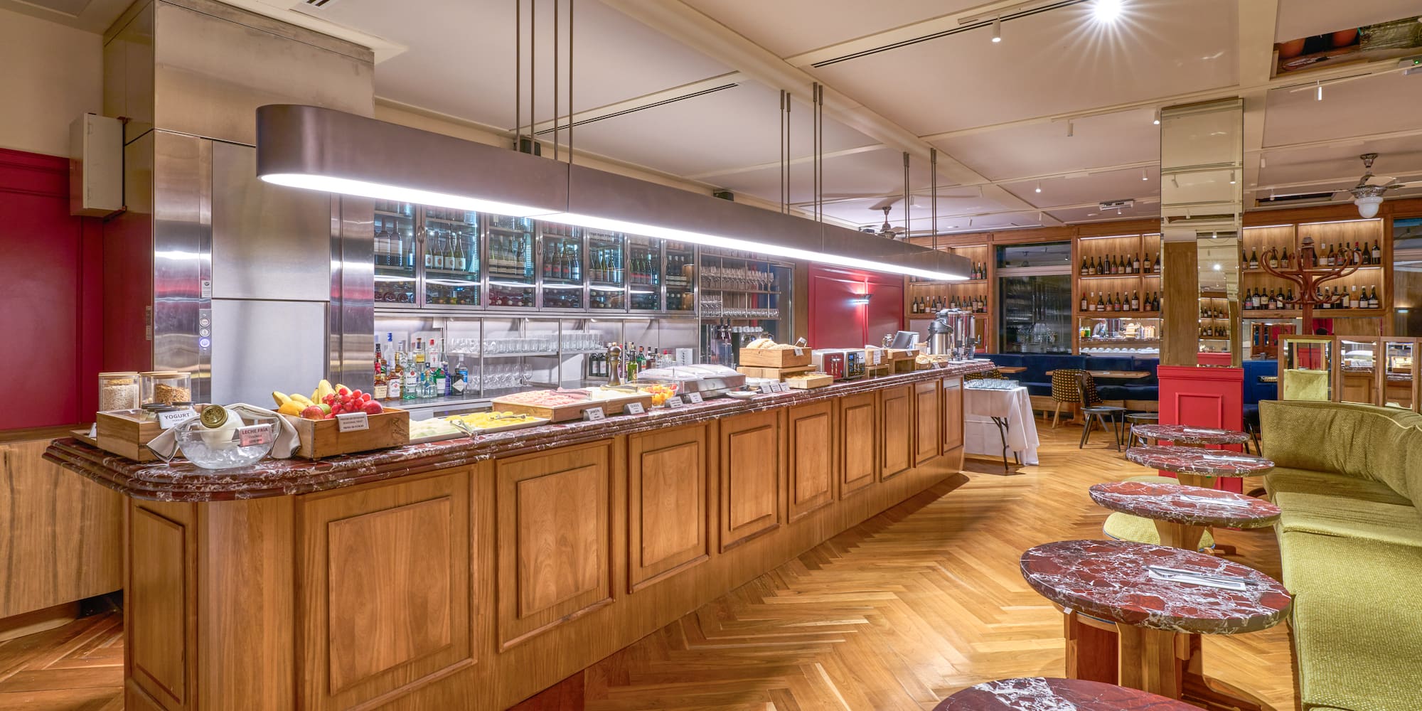 Modern hotel buffet bar with breakfast, wine display, and herringbone wood floor.