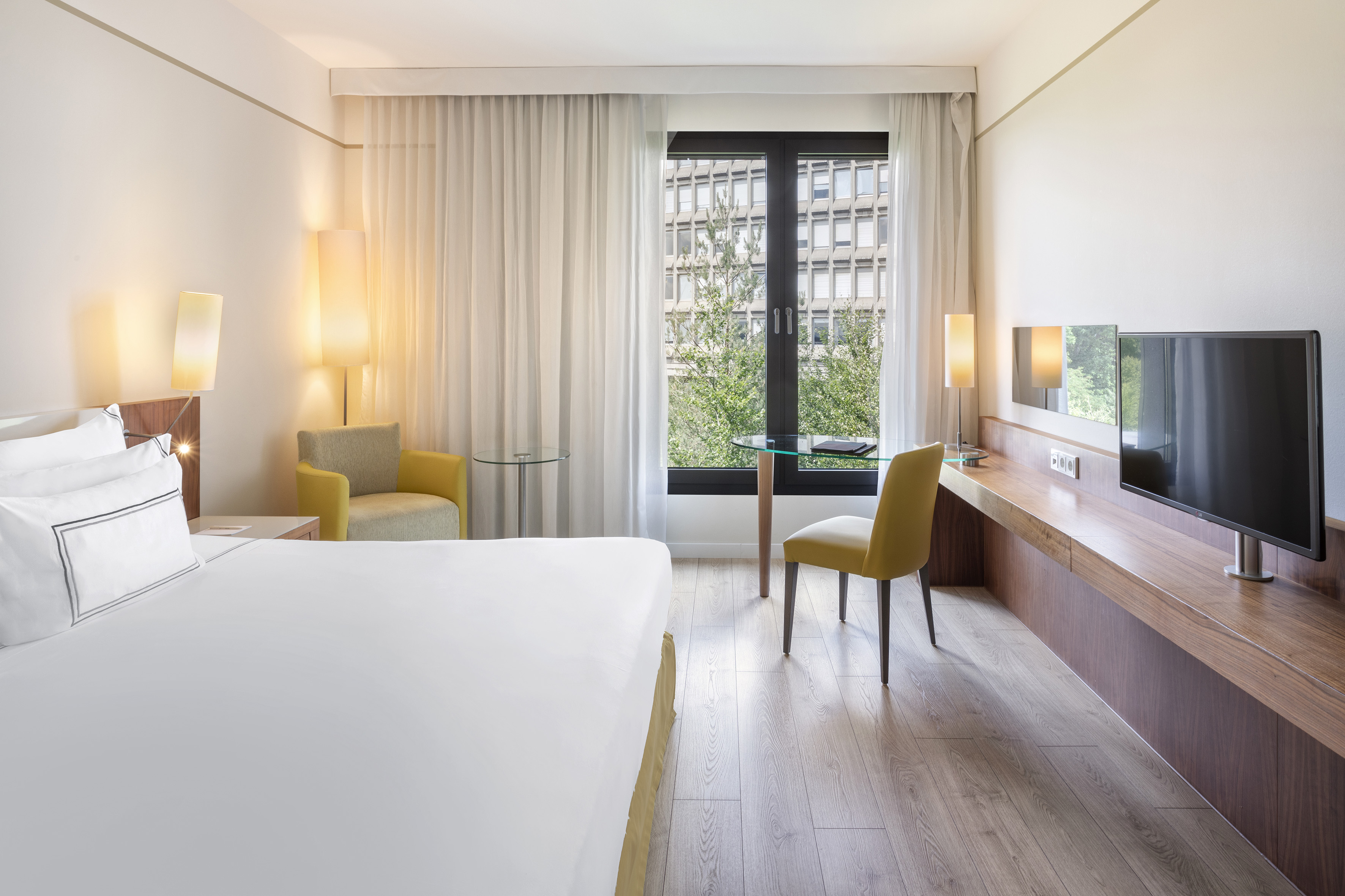 Modern hotel room with bed, work desk, TV, and bright window with city view.