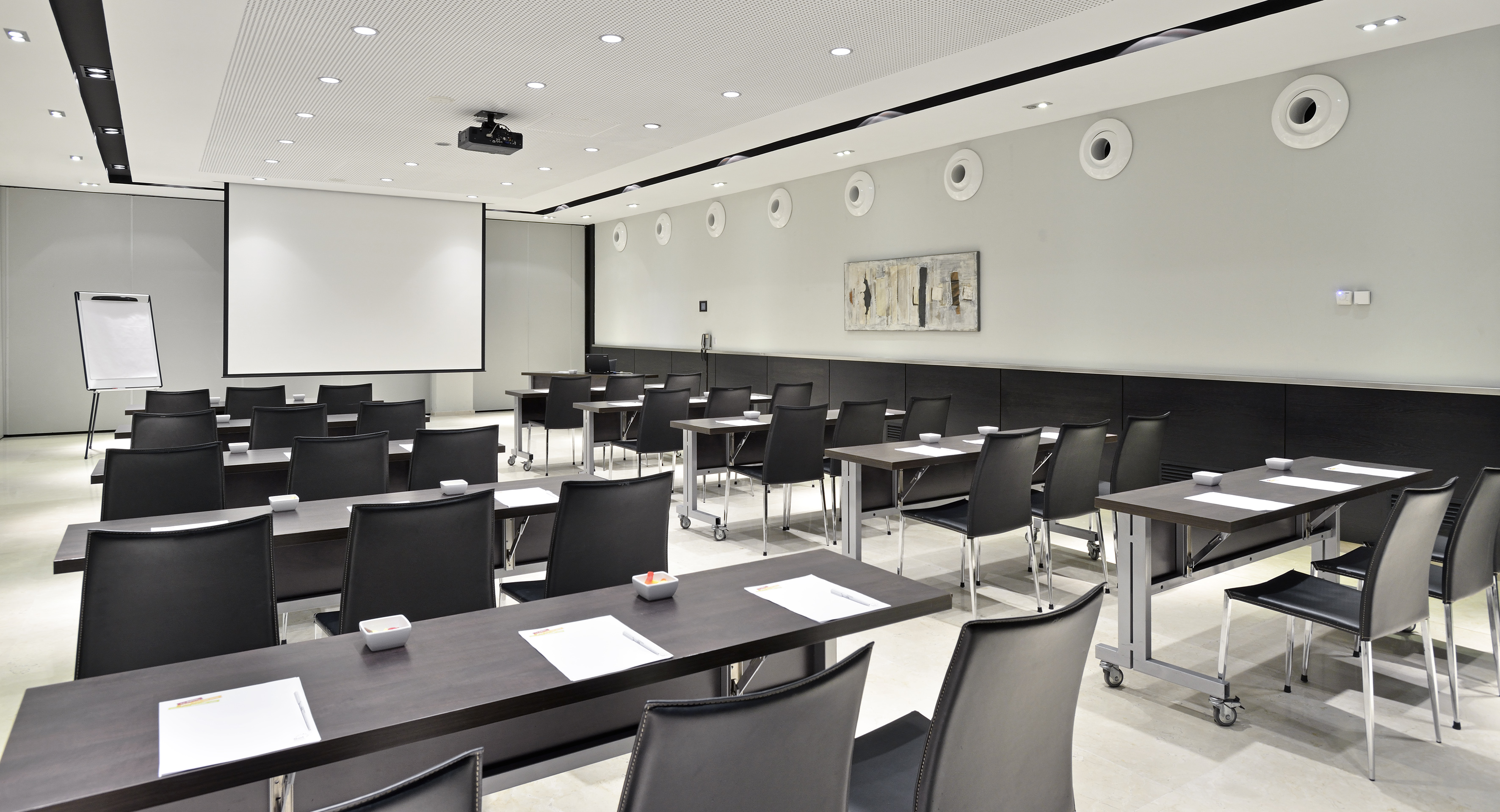 Modern conference room with projector screen, dark tables, and black chairs.