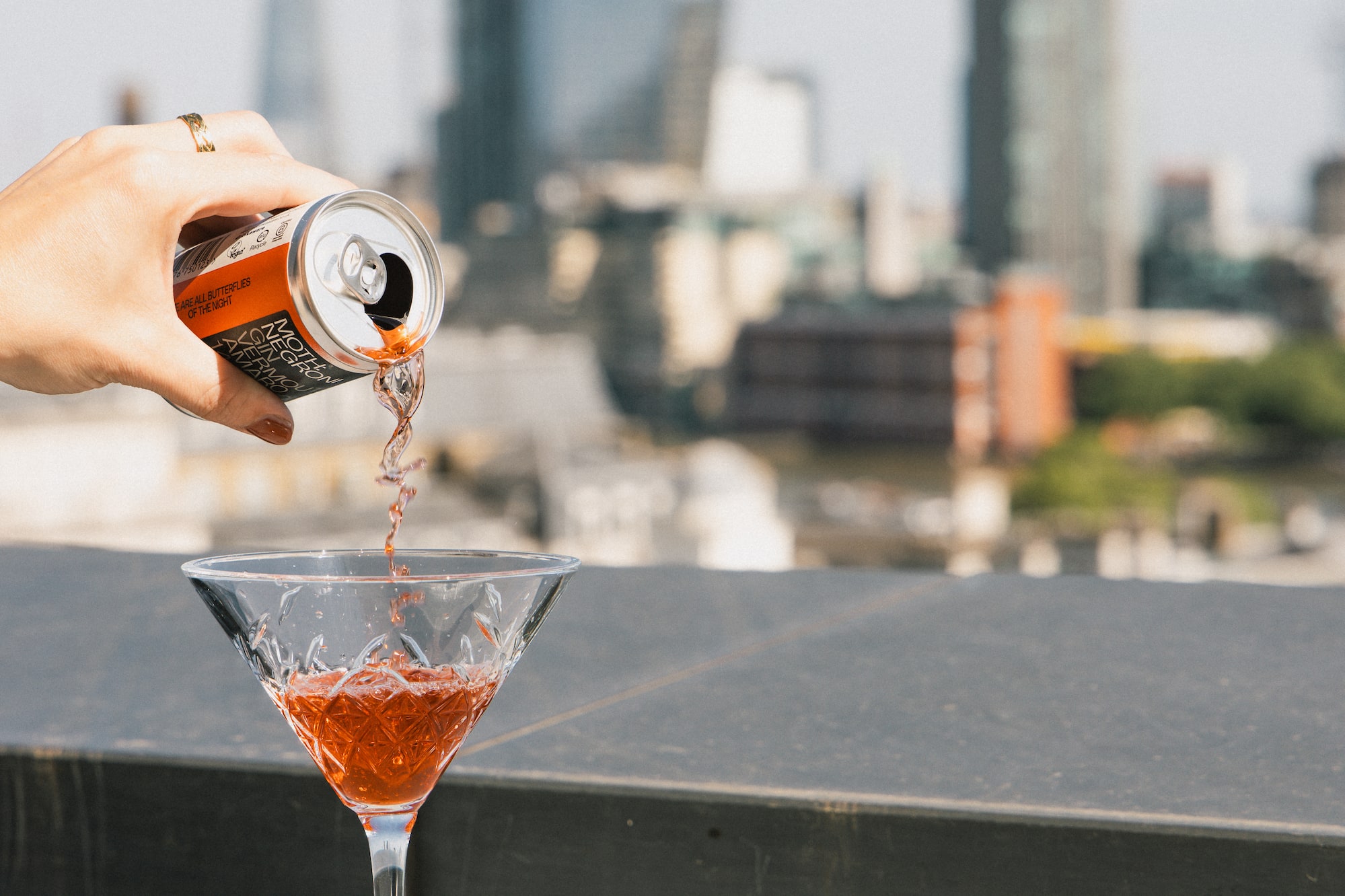 Hand pouring Negroni cocktail into glass, tiramisu, city skyline view.