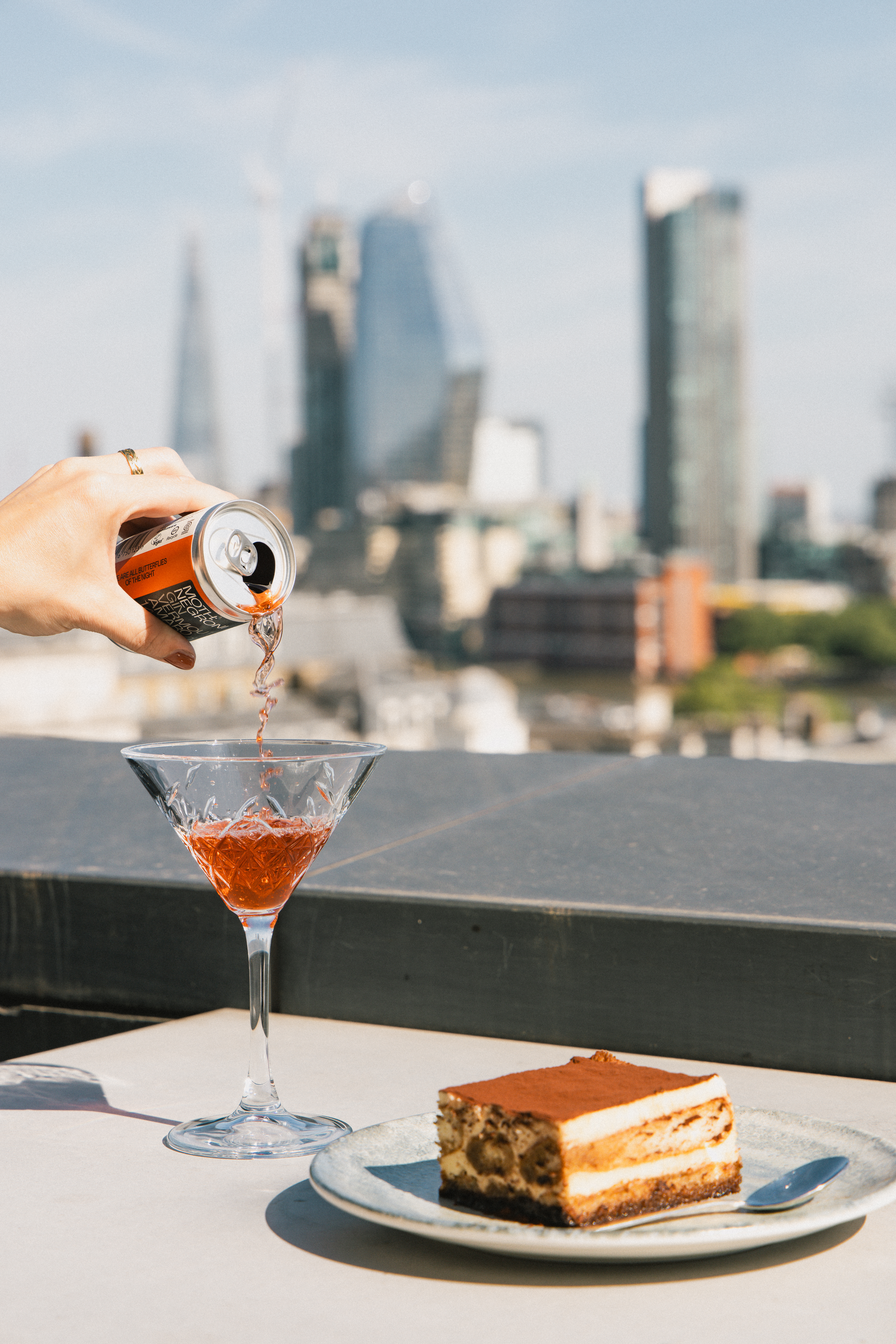Hand pouring Negroni cocktail into glass, tiramisu, city skyline view.