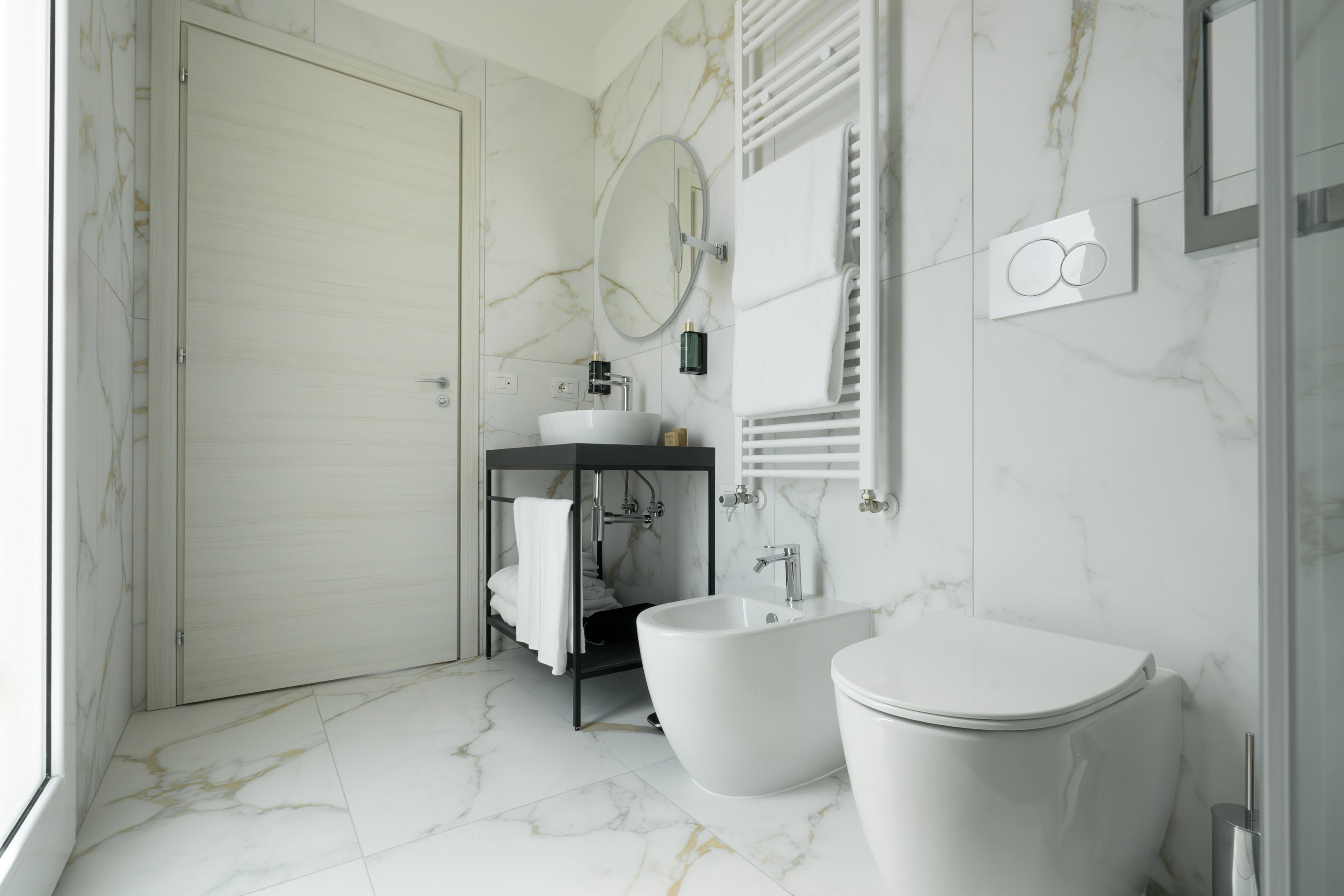 Modern white marble bathroom with toilet, bidet, vanity, and towel warmer.