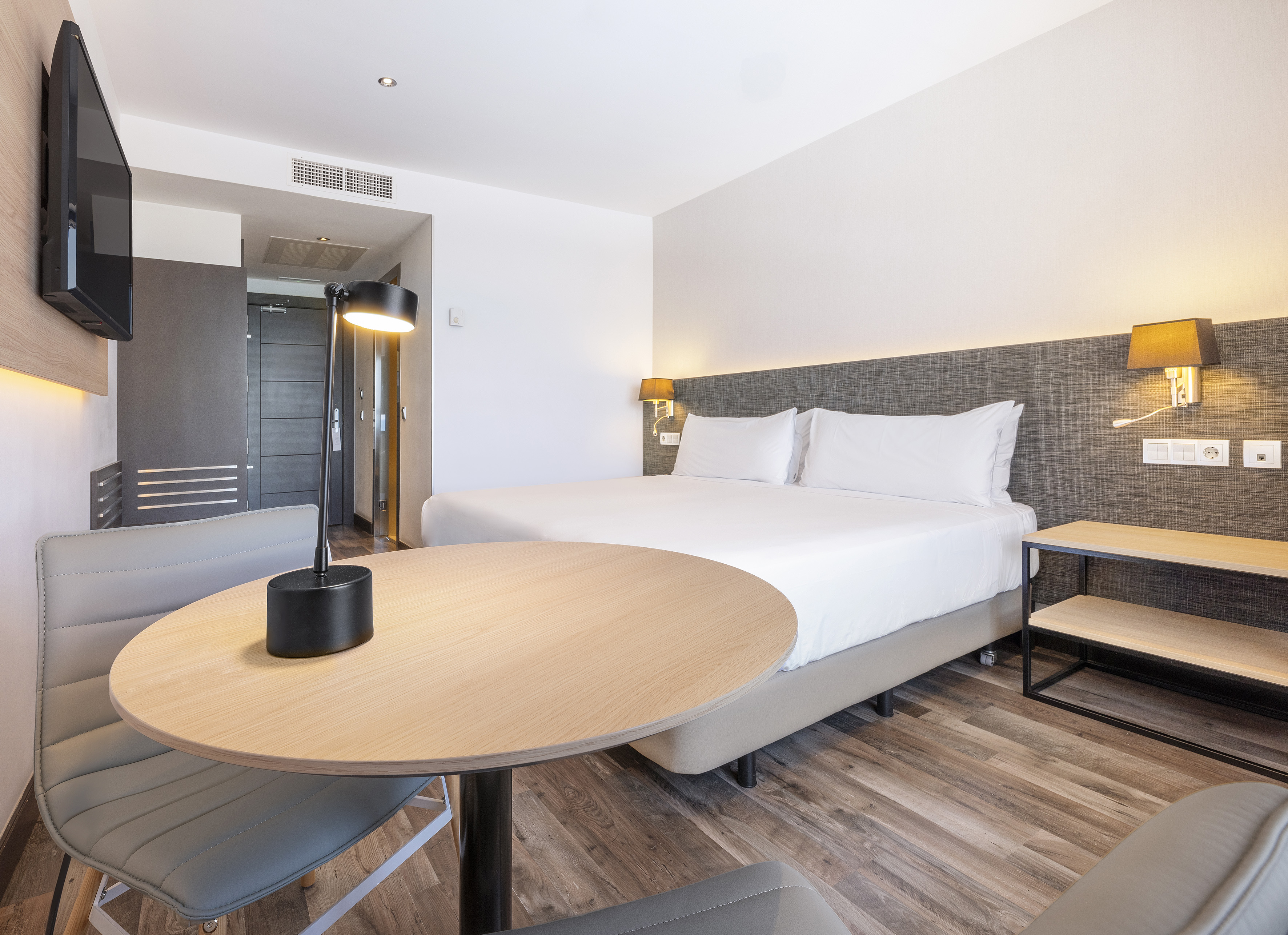 Modern hotel room with large bed, wooden floor, round table, and wall-mounted TV.