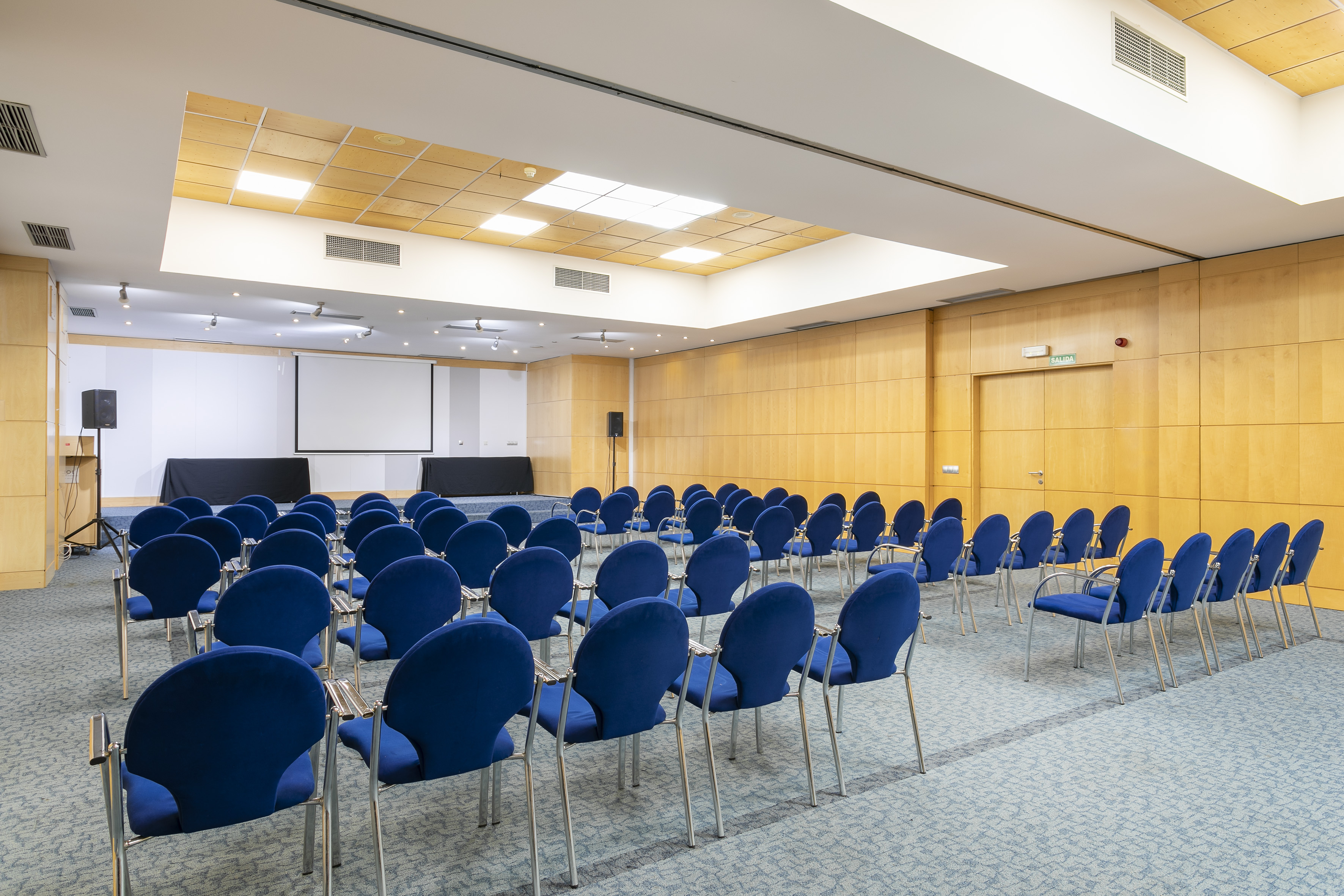 Empty conference room with blue chairs, projection screen, and stage.