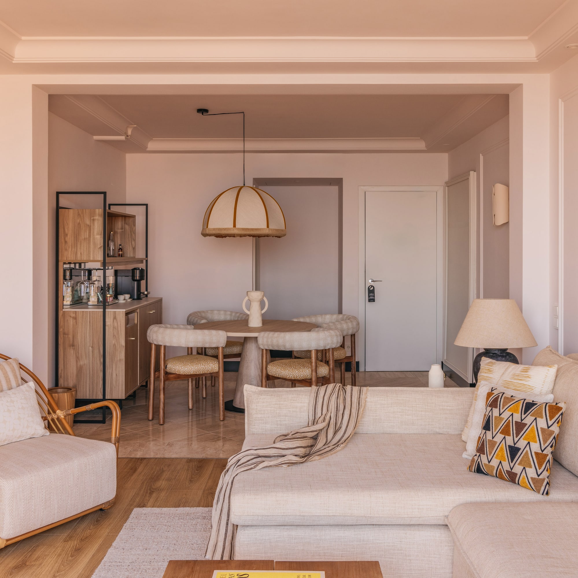 Modern hotel suite living and dining area with neutral decor and rattan chair.
