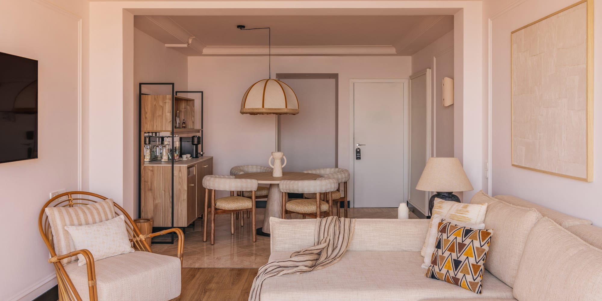Modern hotel suite living and dining area with neutral decor and rattan chair.