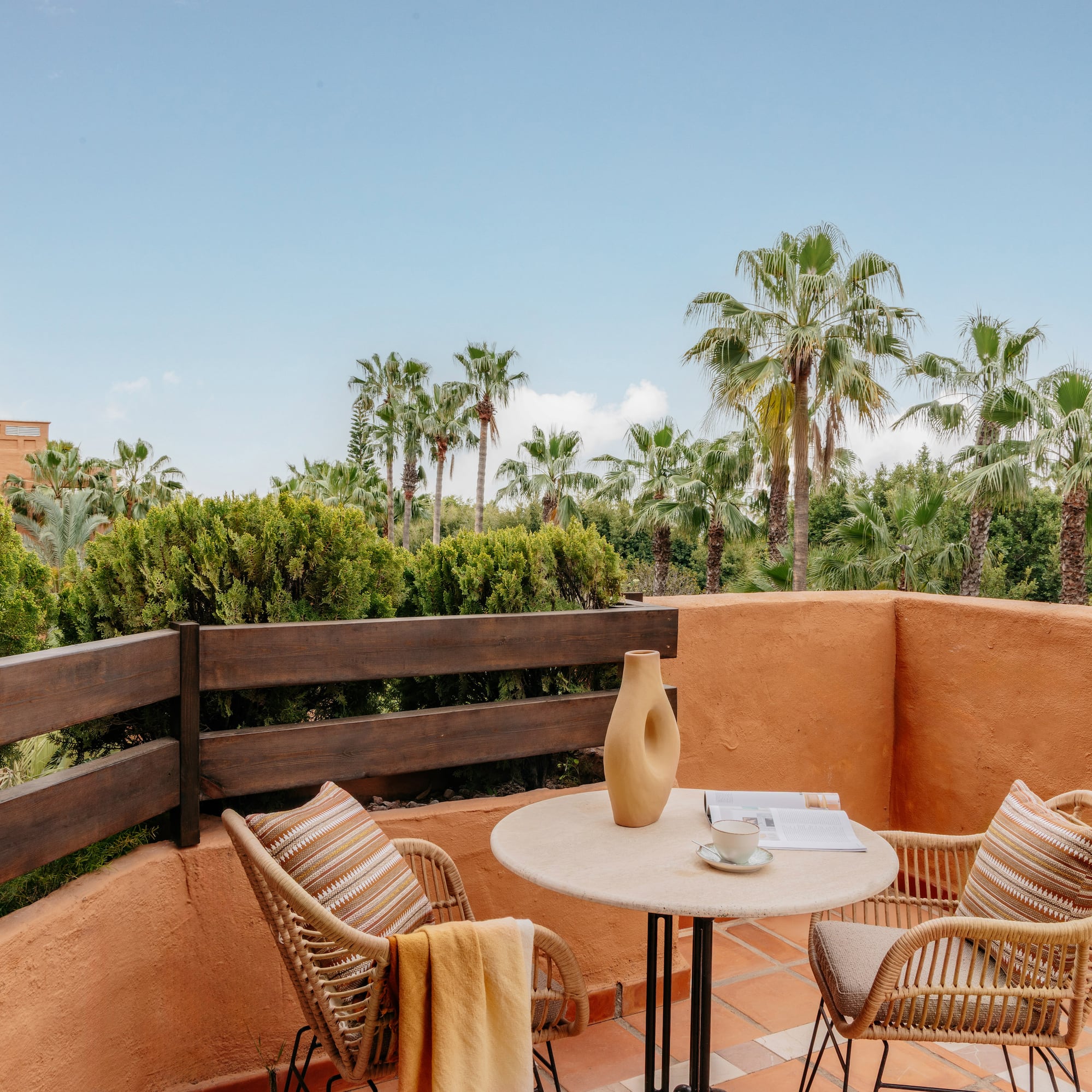 Terracotta balcony with wicker furniture, coffee, and tropical palm trees.