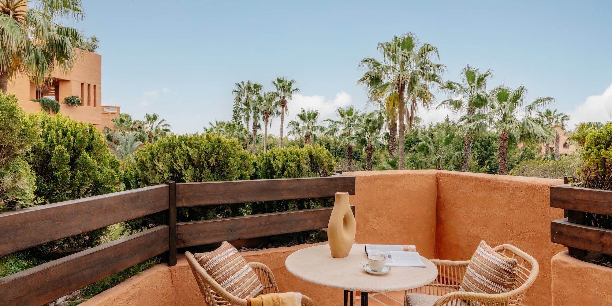 Terracotta balcony with wicker furniture, coffee, and tropical palm trees.